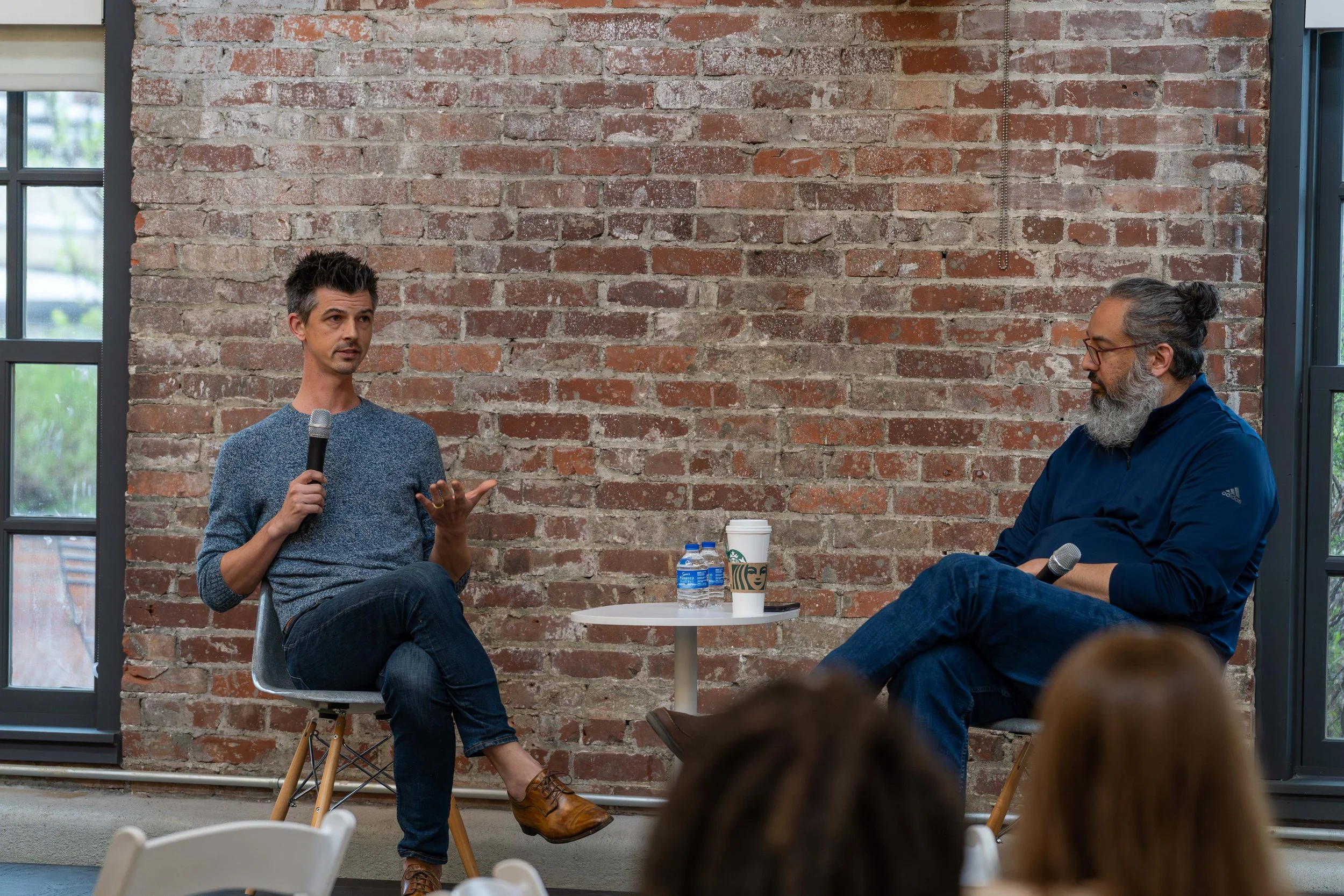 A man speaking into a microphone while seated and a man listening in a discussion setting with a brick wall background and windows.
