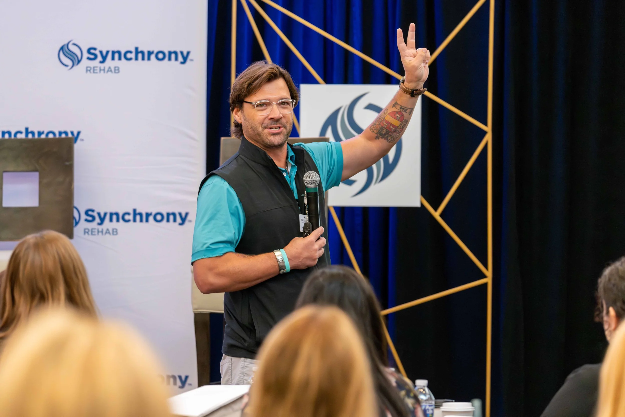 A man with glasses and tattoos speaking into a microphone during a presentation at a conference, with a backdrop displaying the logos of 'Synchrony REHAB' and a stylized swirl design.