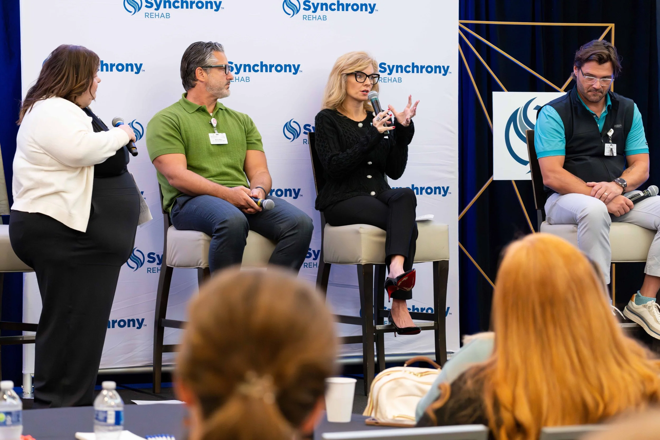 A panel discussion at Synchrony Rehab with four individuals on stage, including two women and two men, with a logo backdrop. One woman is speaking into a microphone, while the others listen.