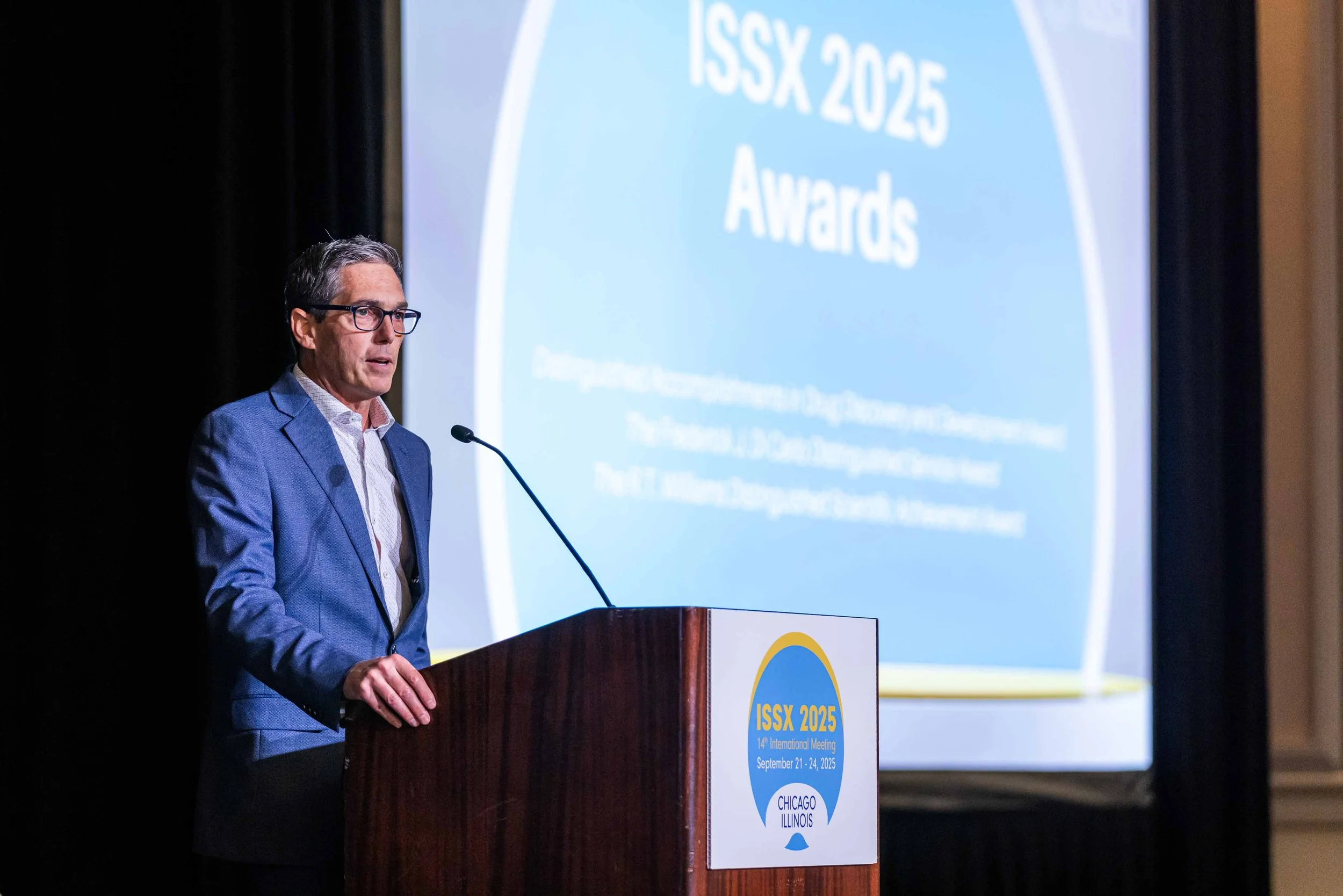 A man in a blue suit and glasses speaks at a podium during the ISSX 2025 awards ceremony, with a large screen displaying the event details behind him.