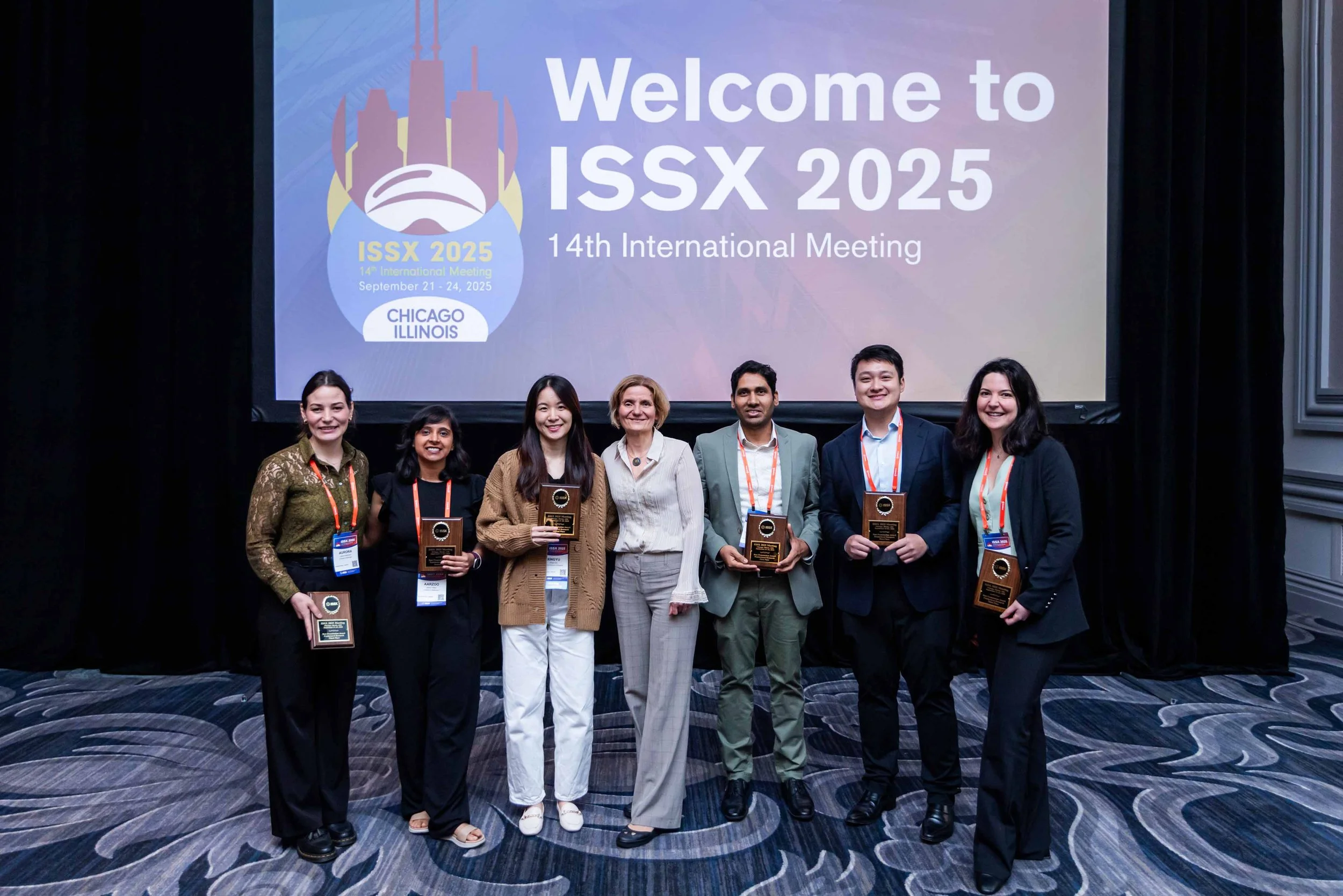 Group of seven people standing in front of a large screen with a welcome message for ISSX 2025, holding awards during the 14th International Meeting in Chicago, Illinois.
