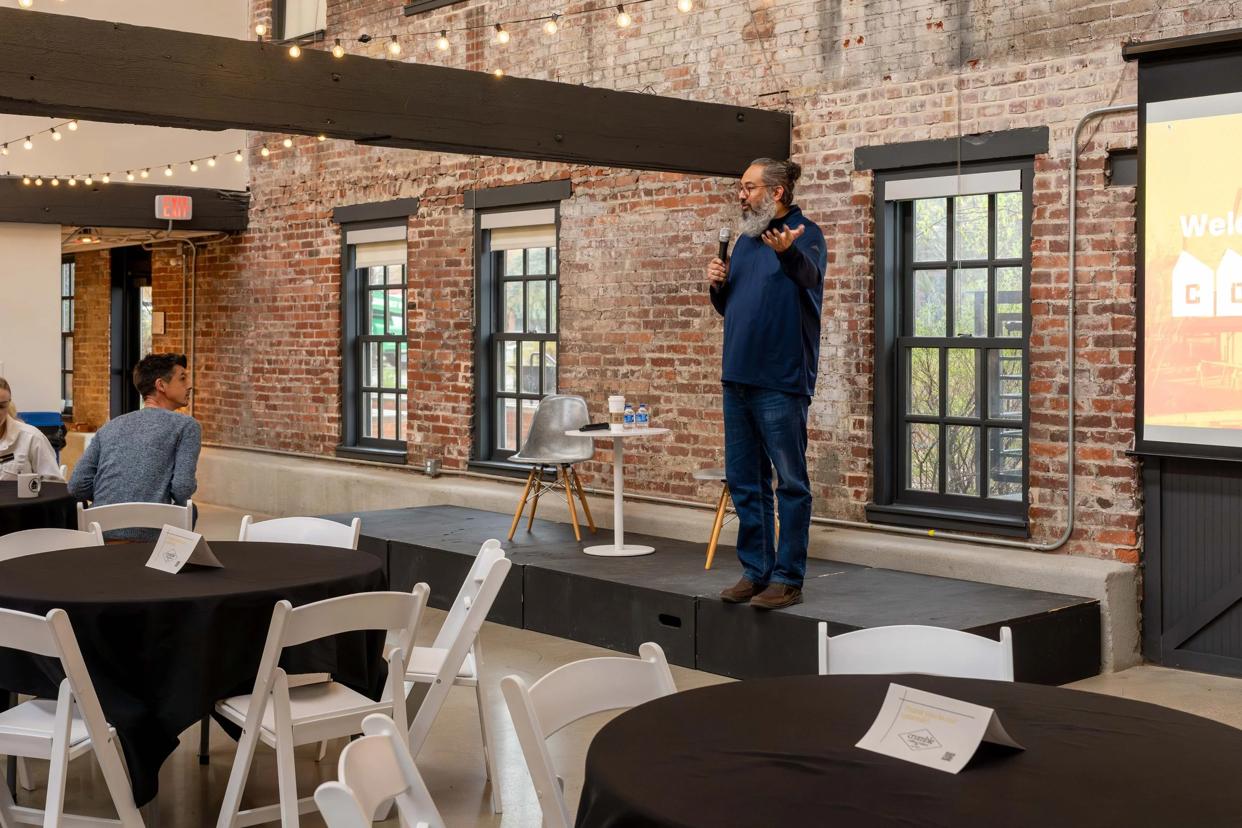A man with gray hair and a beard, wearing a navy jacket and jeans, speaking into a microphone on a small stage in a brick-walled room with string lights overhead. There are a few tables with white chairs and black tablecloths, and a presentation scre