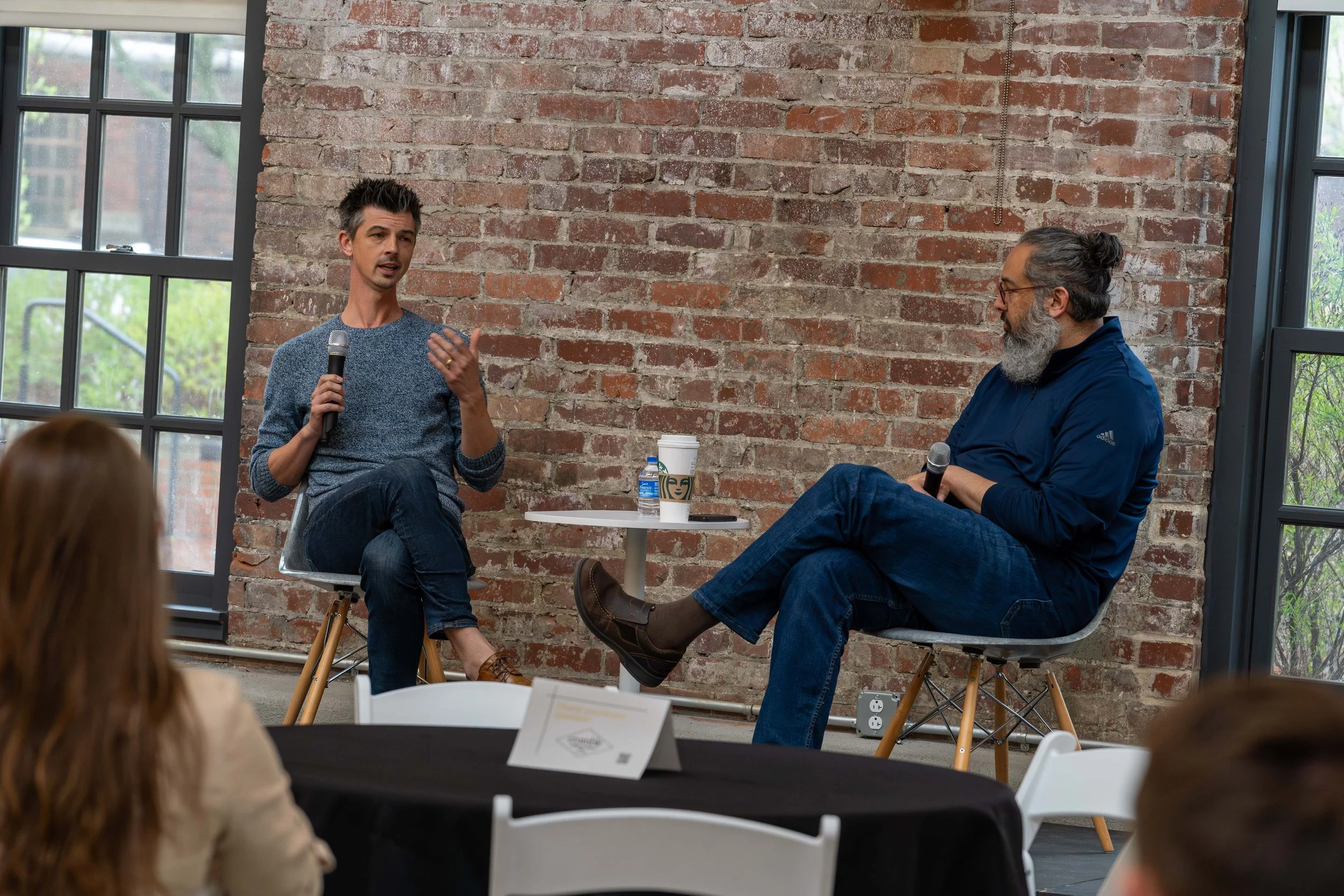 Two men sitting on chairs engaged in a discussion, holding microphones, in a room with a brick wall and large windows.