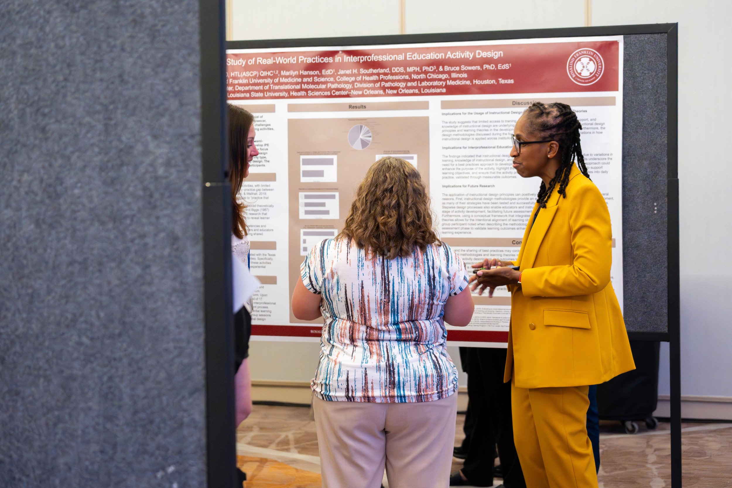 A woman in a yellow blazer and glasses discusses a research poster with two other women, one with curly hair and in a colorful top, at an academic conference.
