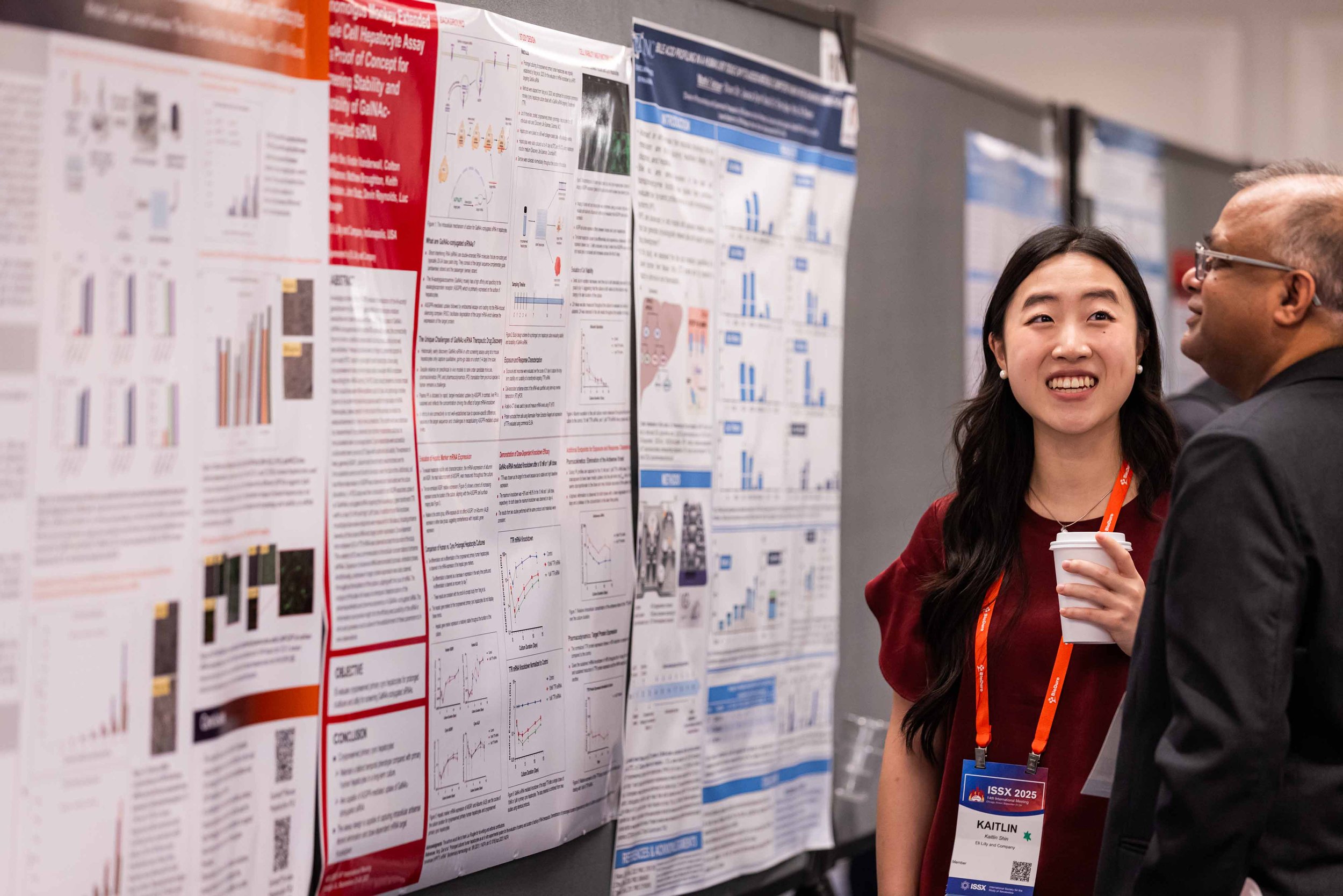 Two people, a woman and a man, are standing in front of research posters at a conference. The woman is smiling, holding a coffee cup, and wearing a name badge. The background consists of scientific posters with charts and graphs.