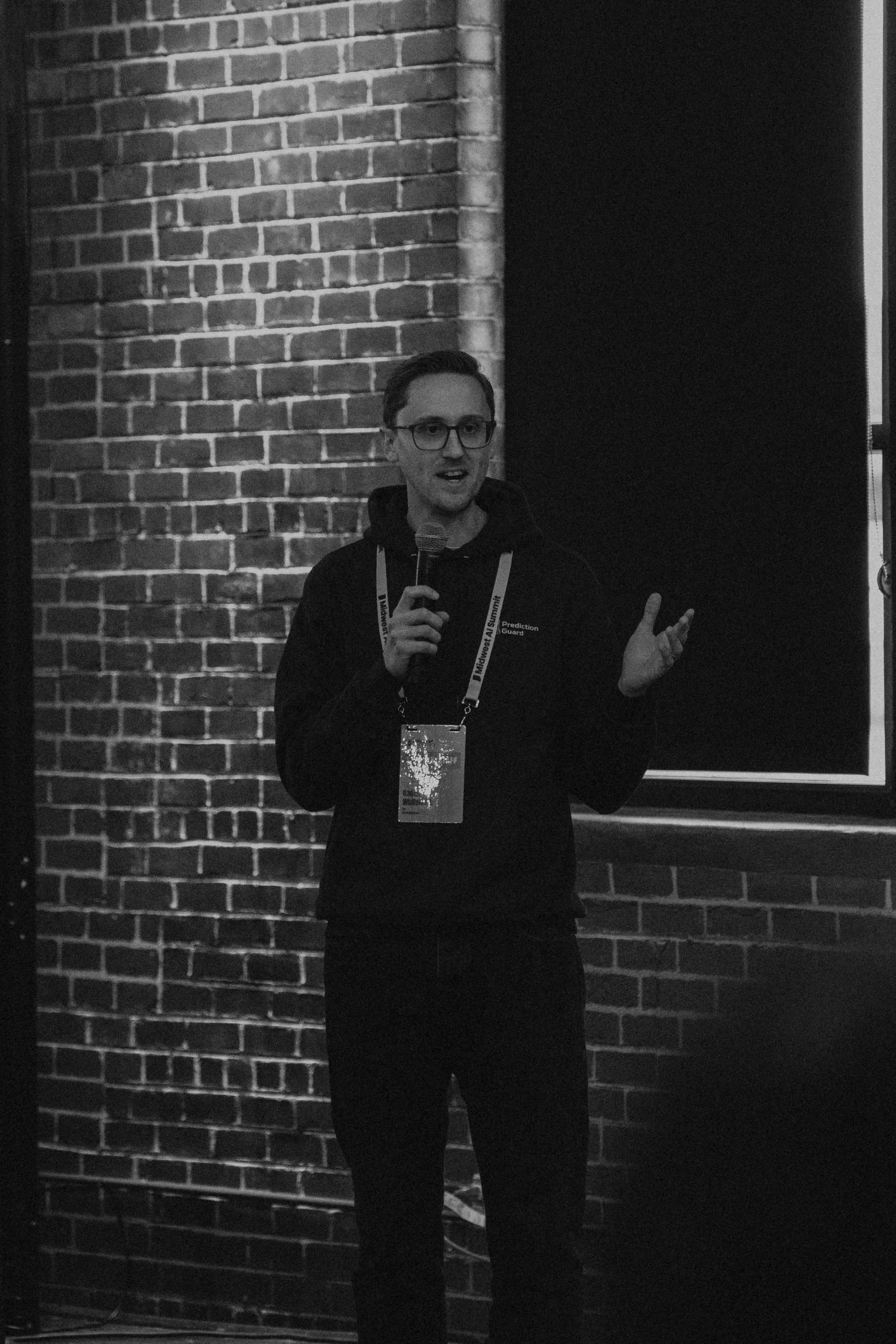 A young man with glasses holding a microphone, speaking in front of a brick wall during a presentation or event.