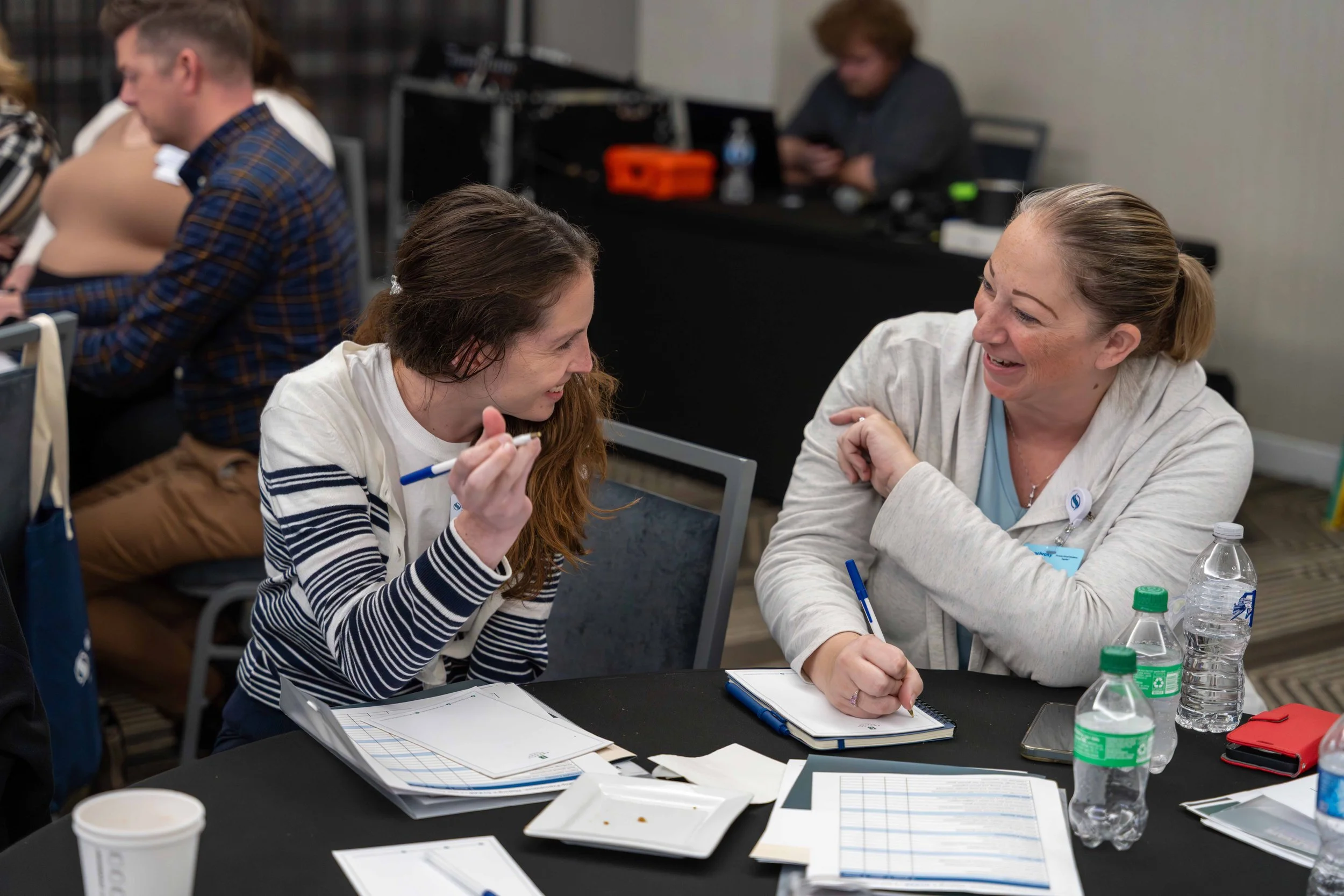 Two women are smiling and talking at a conference table, with notebooks and water bottles in front of them. One is holding a pen, and they appear engaged in a conversation.