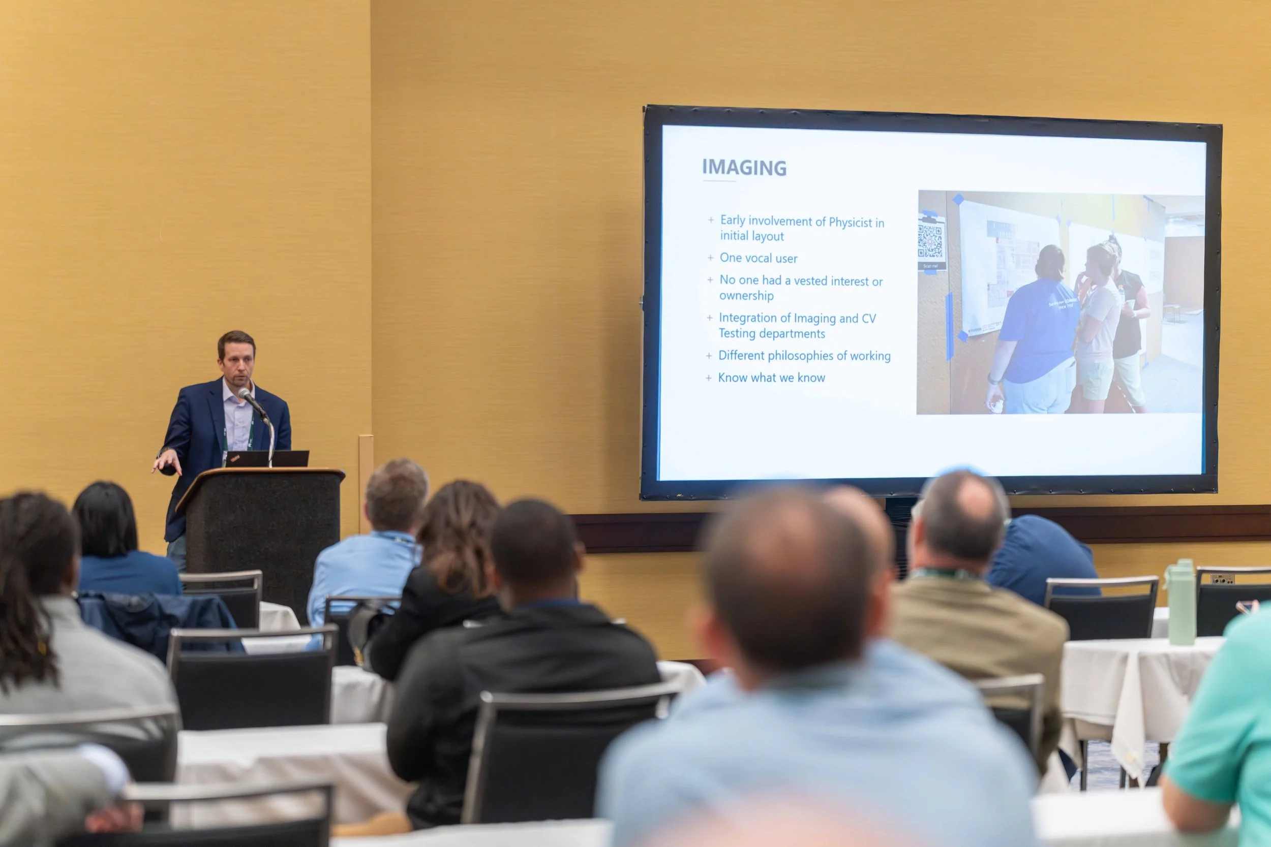 A man in a blue suit giving a presentation at a conference with a large screen displaying a slide titled 'IMAGING'. The slide contains bullet points and a photo of people viewing posters.