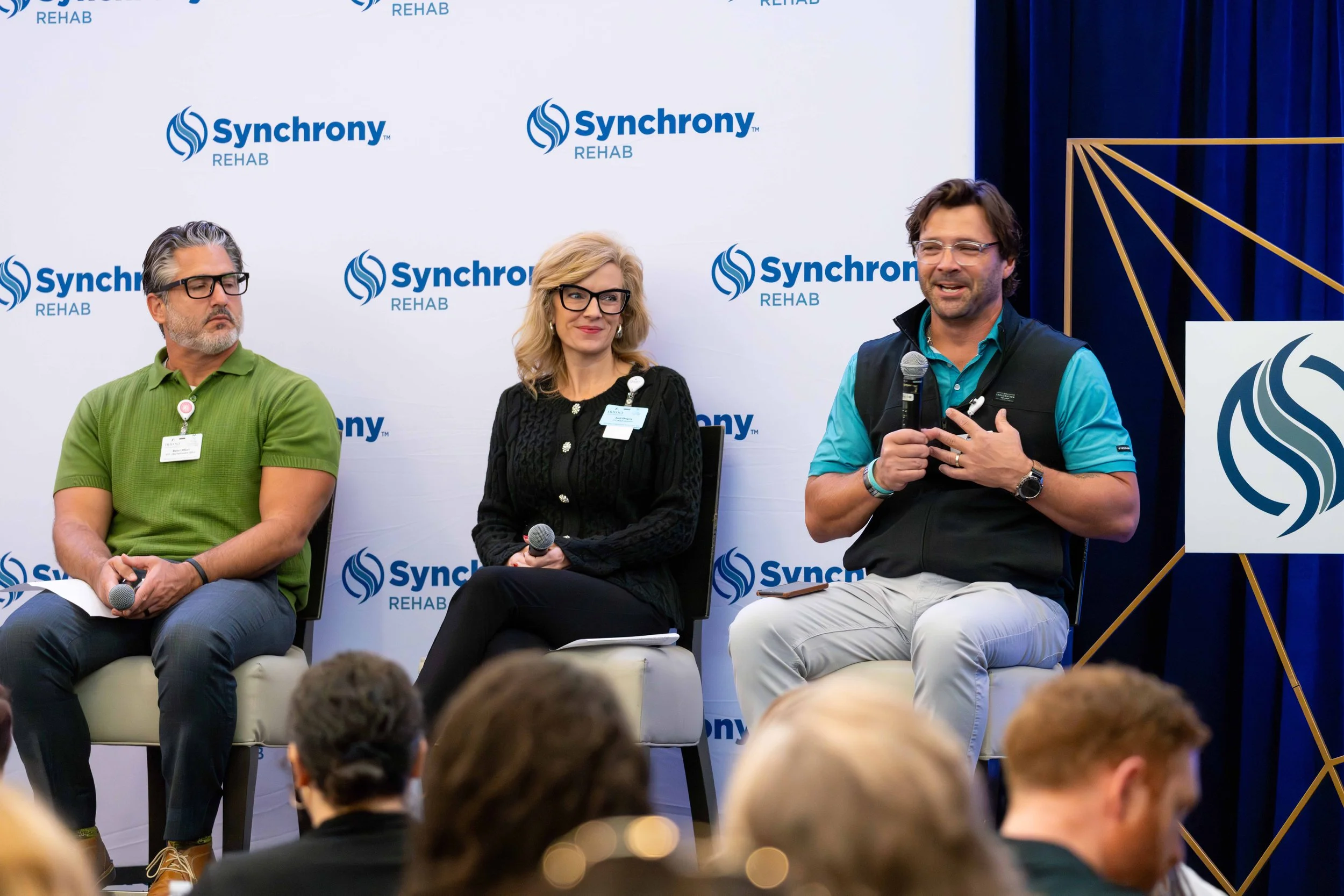 Three people sitting on a stage at a Syncory Rehab event, with a white backdrop displaying the company's logo. The person on the left is a man wearing a green polo shirt and glasses, the woman in the middle is wearing a black sweater and glasses, hol