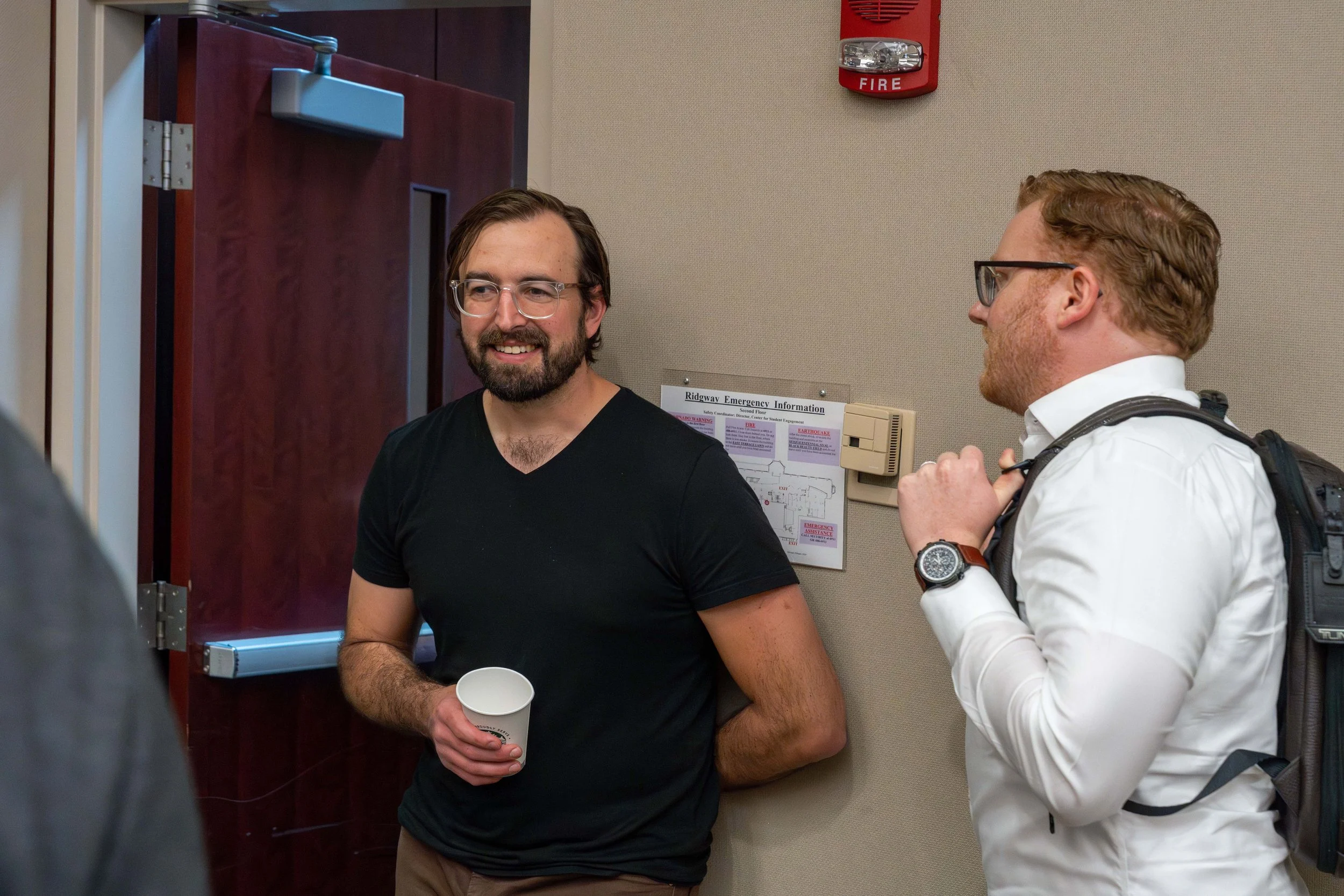 Two men engaged in conversation at a meeting or social gathering, standing indoors near a door and an emergency information sign on the wall.