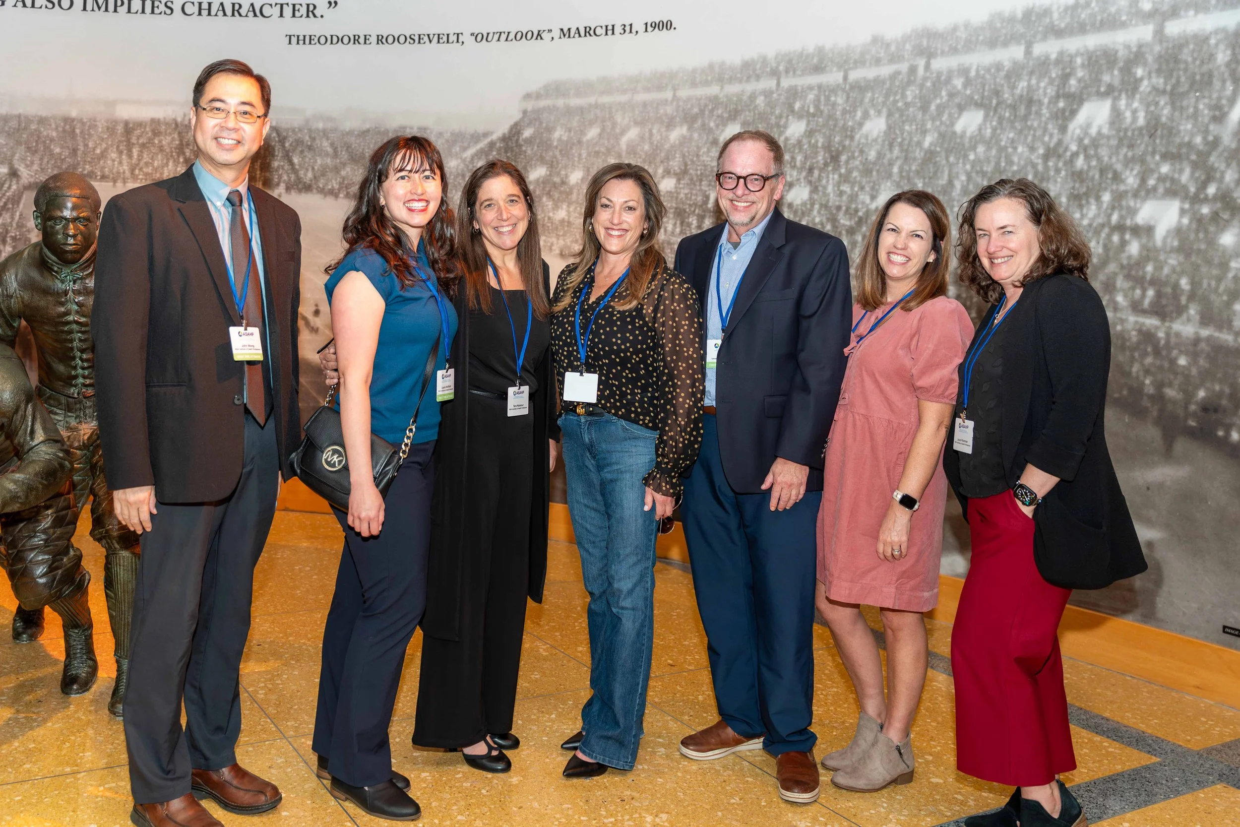 Group of eight diverse adults, six women and two men, standing together at an indoor event, smiling for the camera. They are wearing business casual attire with blue conference lanyards. A large historical photograph or mural is seen in the backgroun