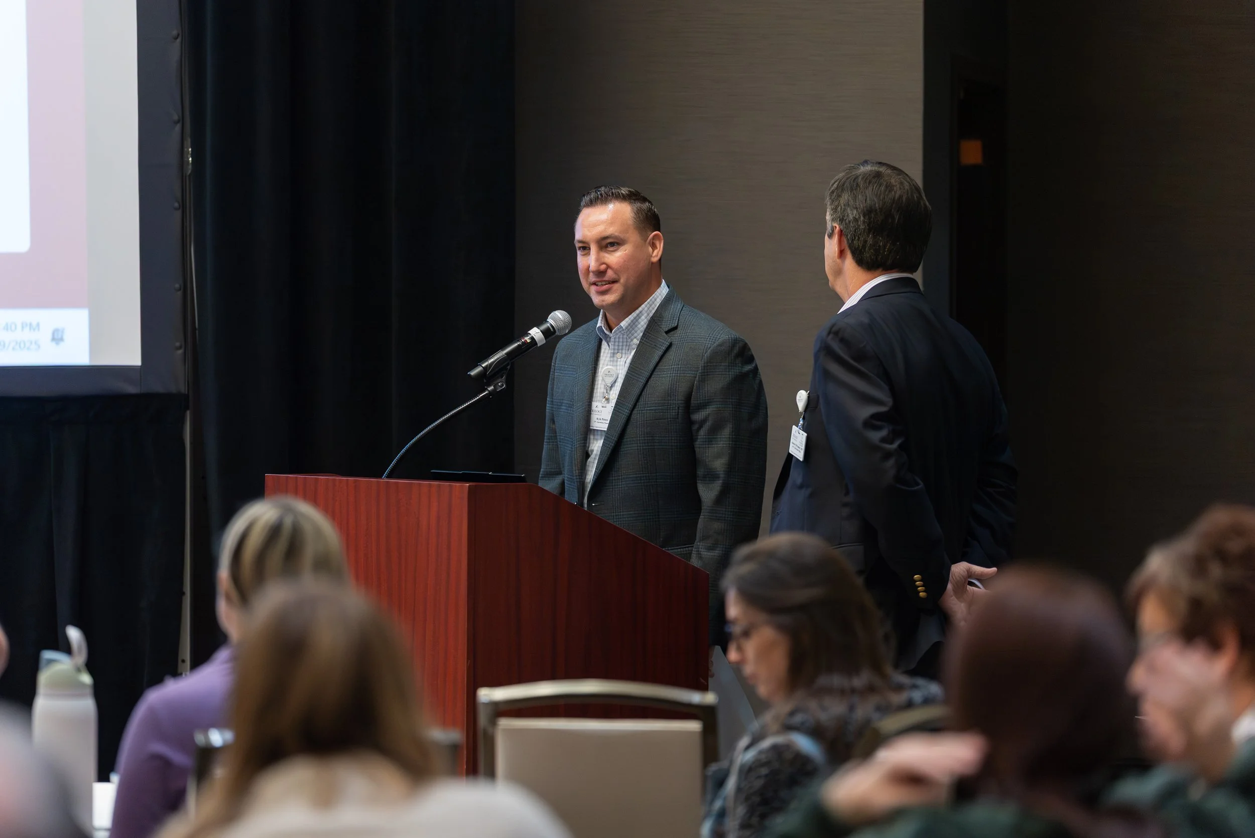 A man in a gray suit speaks at a podium during a conference, with another man in a dark suit standing nearby. An audience listens in the foreground.