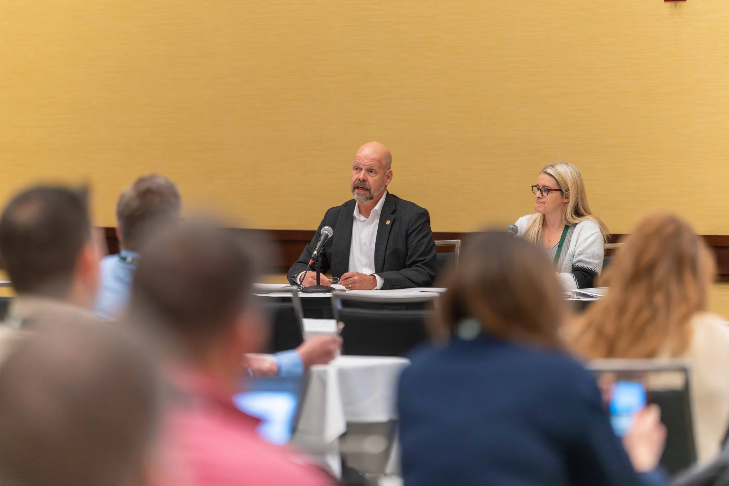 A man and woman seated at a panel during a conference, with an audience in front of them. The man is speaking into a microphone, wearing a black blazer and white shirt, while the woman is listening with a smile, wearing glasses and a light-colored sw