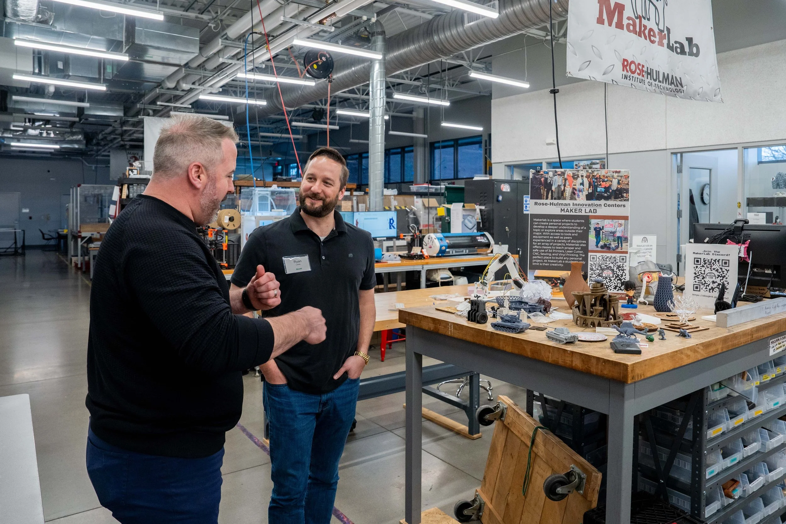 Two men are talking next to a display table with various small 3D printed objects inside a modern manufacturing or innovation lab, with industrial ceiling pipes and a banner that reads "Maker Lab."
