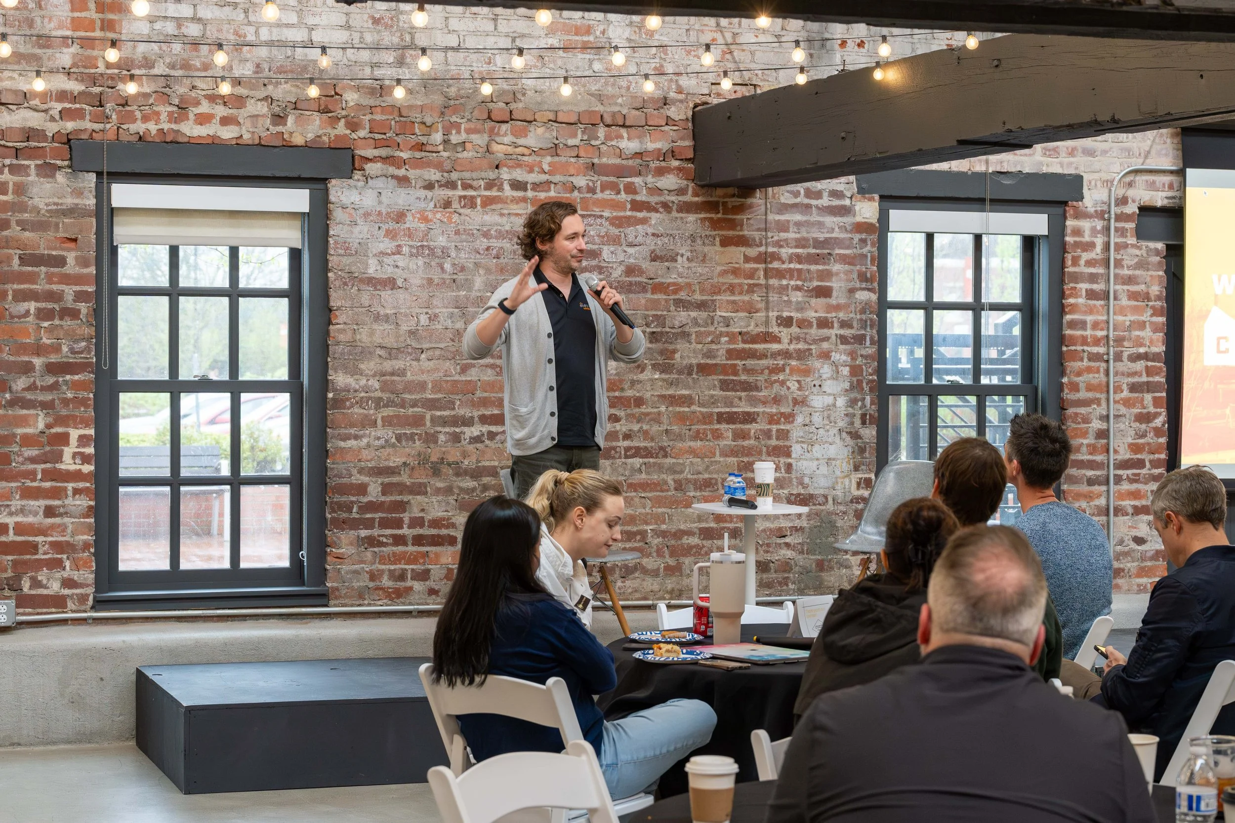 A man giving a presentation to a group of people in a room with brick walls and large windows, holding a microphone and gesturing with his hand. The audience is seated at tables with coffee cups and notebooks.