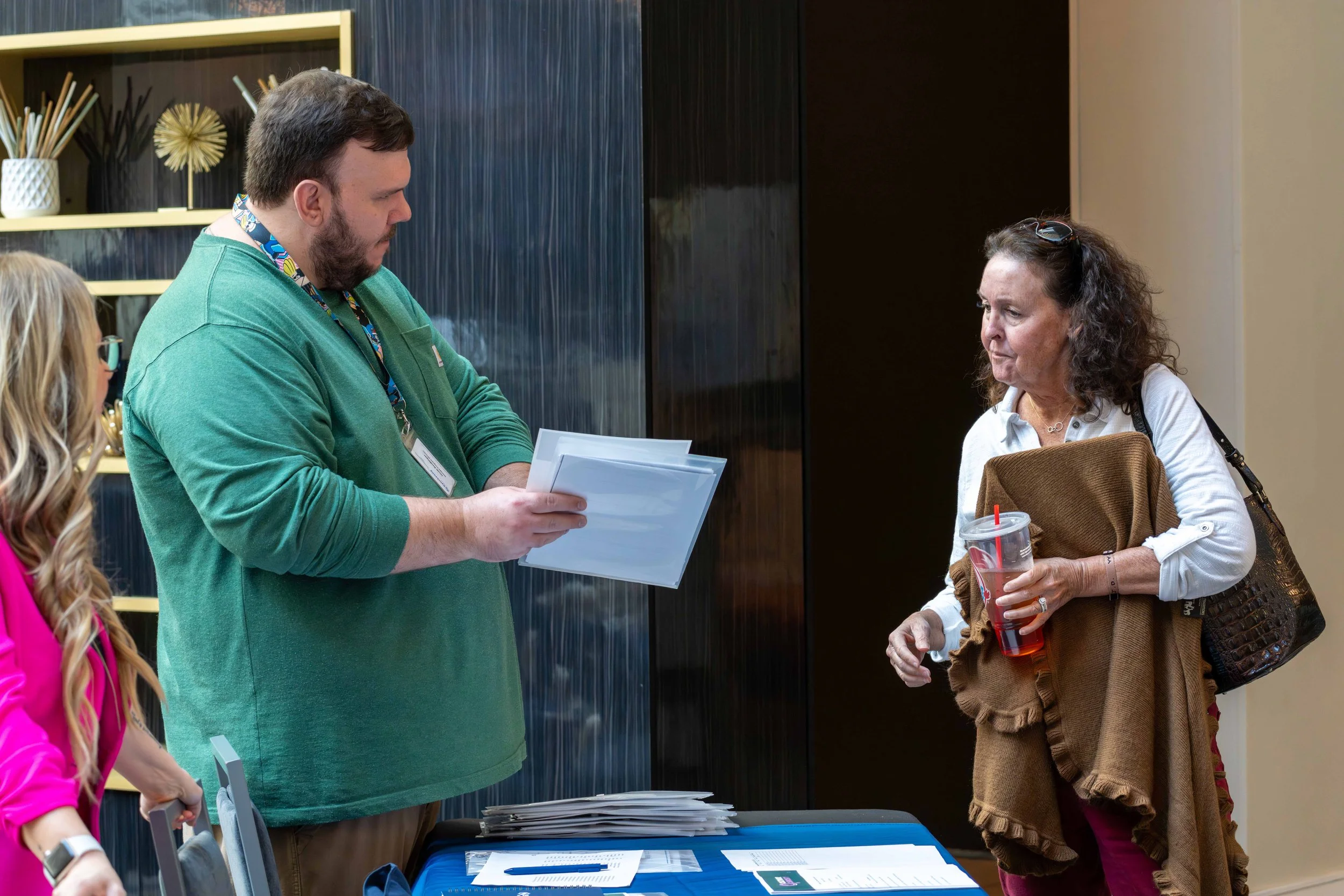 A woman with dark curly hair holding a cup with a red straw and a brown jacket, talking to a man with a beard and a green shirt who is examining paperwork at a table.