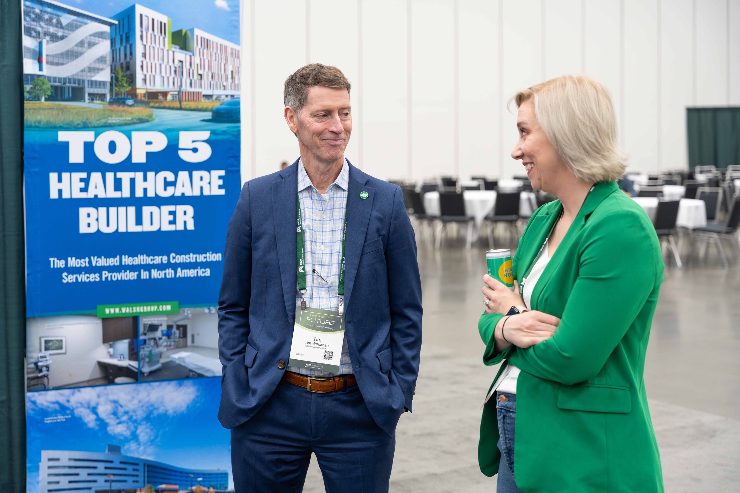 A man and woman having a conversation at a conference, standing next to a large poster that reads "Top 5 Healthcare Builder" with a building and healthcare images in the background.