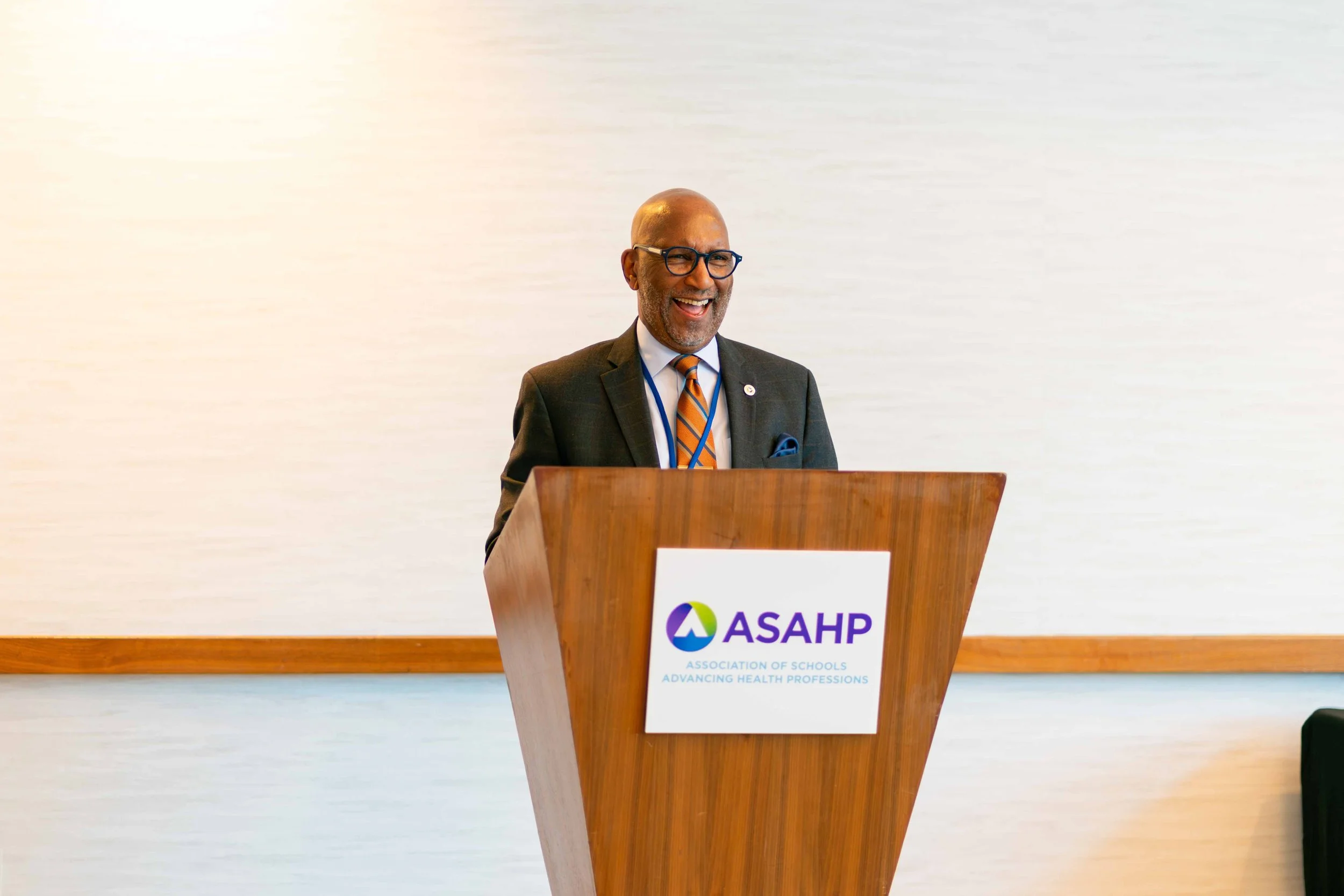 A smiling man in a suit and tie standing at a podium with the ASAPH logo, delivering a speech.