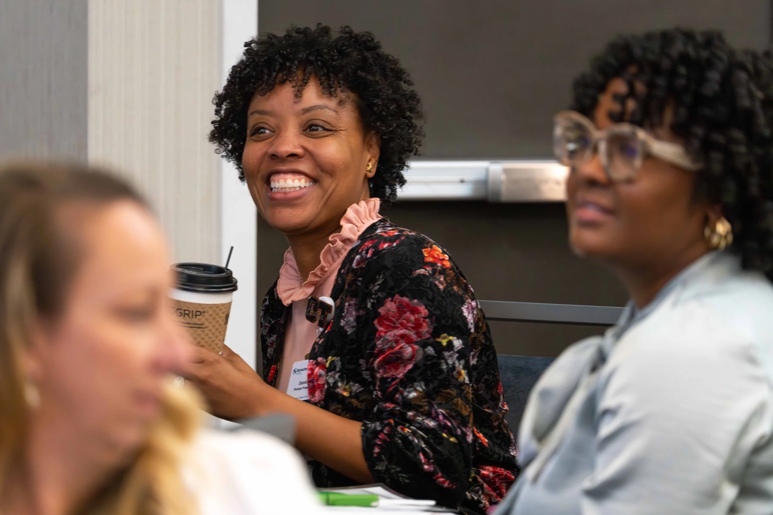 Three women at a meeting, with one woman smiling and holding a coffee cup, wearing a floral jacket, and the other two women partly visible.