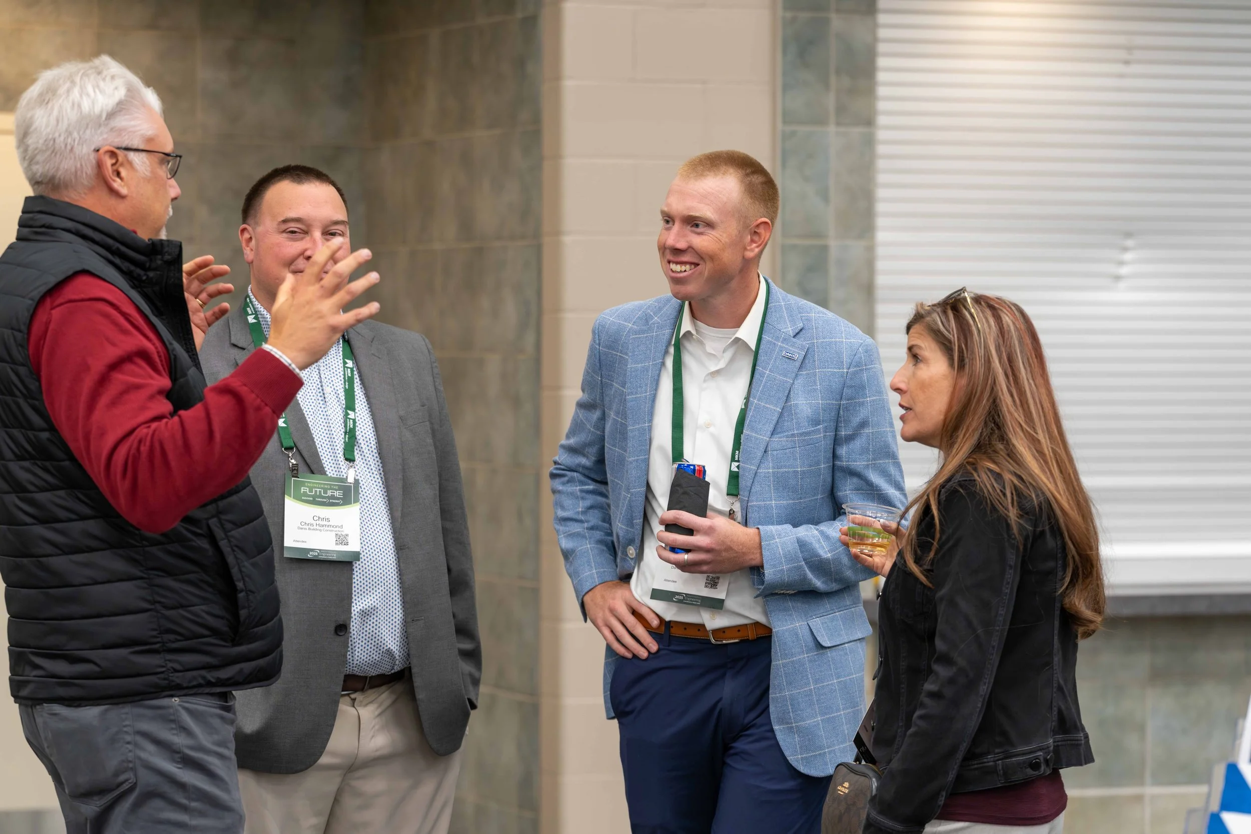 Four people engaged in conversation at a professional event, standing in a lobby or conference area, with two men wearing suits and a woman in a black jacket, one man wearing glasses and a name badge, and the woman holding a drink.