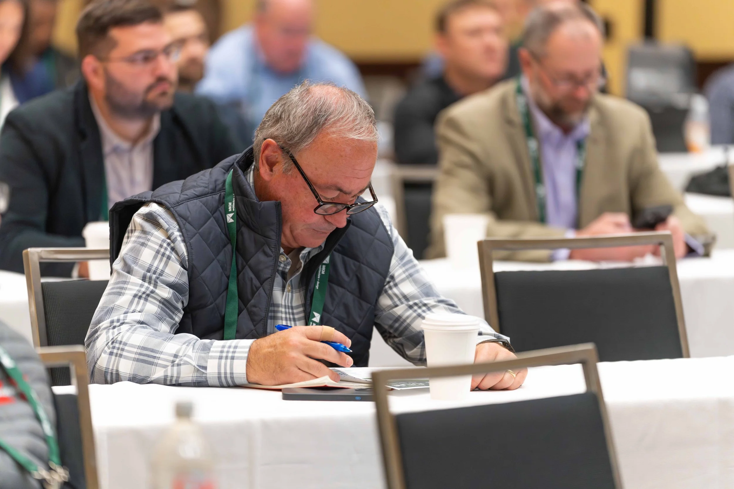 A man with glasses, wearing a plaid shirt and a navy vest, is sitting at a conference table taking notes. He's in a busy conference room with other attendees, some of whom are looking at their phones.