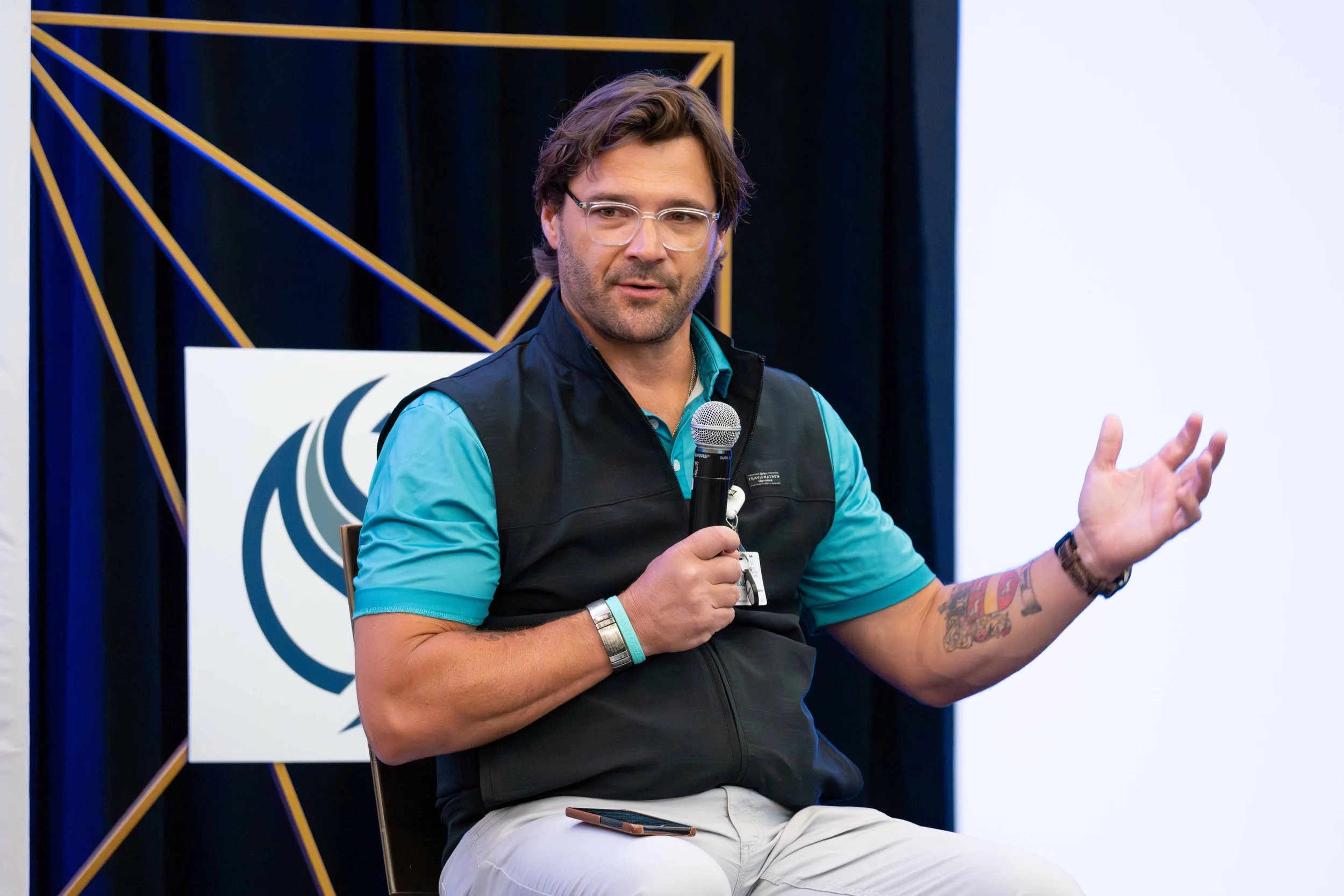 A man with glasses and tattoos on his right arm is sitting on a chair, speaking into a microphone during a presentation or panel discussion. He is wearing a blue shirt and a black vest, with a smartphone on his lap, and gesturing with his left hand.
