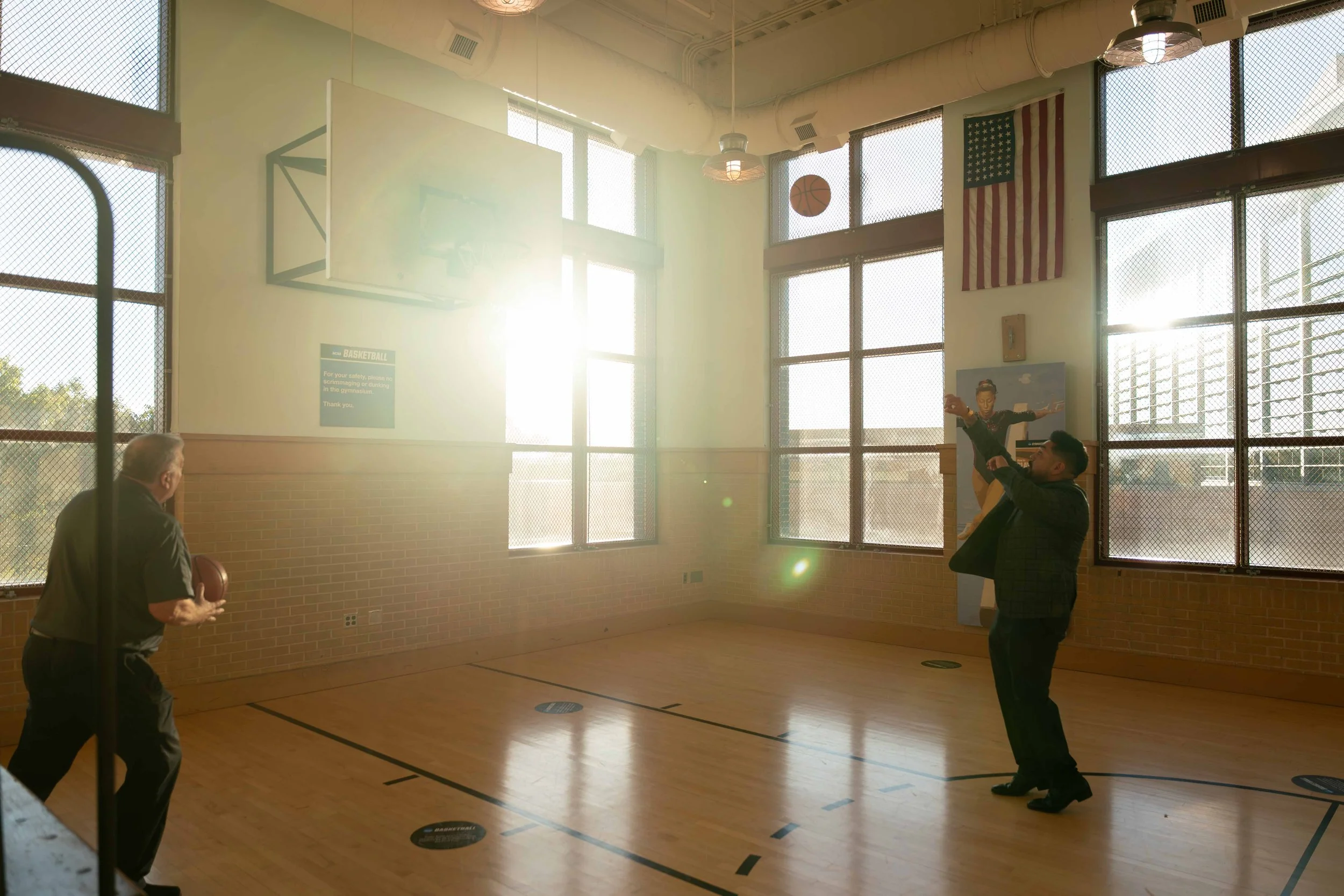 Two men playing basketball in a gym with large windows, American flag, and painting of a basketball player on the wall. Sunlight streaming in through the windows.