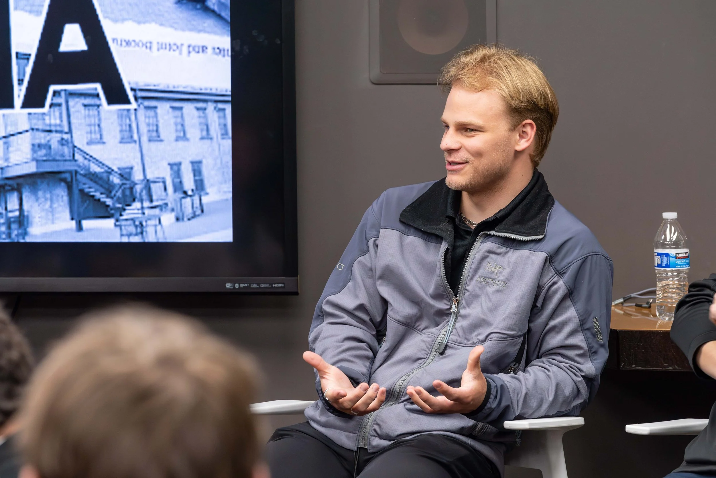 A young man with blonde hair speaking in a classroom or meeting room, seated next to a large screen displaying a building and the letter 'A'.
