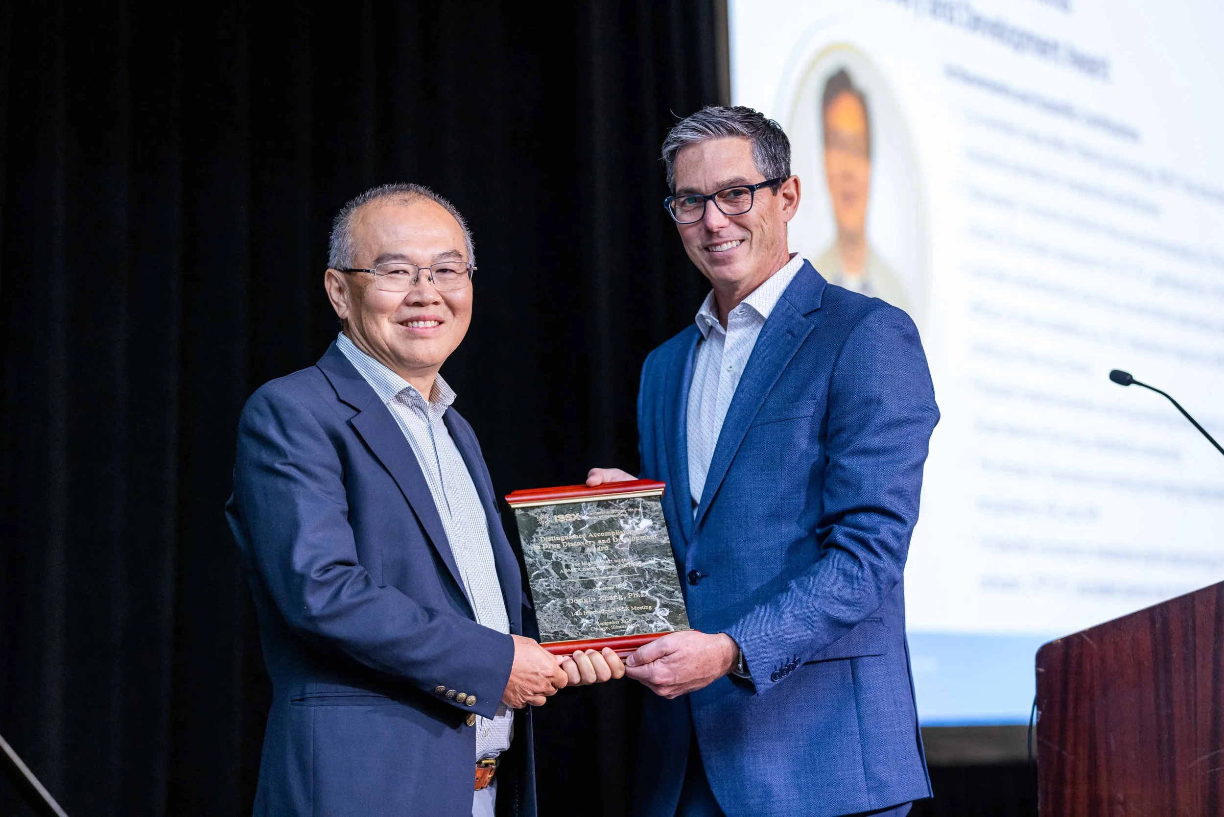 Two men in suits on stage, one presenting an award or plaque to the other, with a presentation slide in the background.
