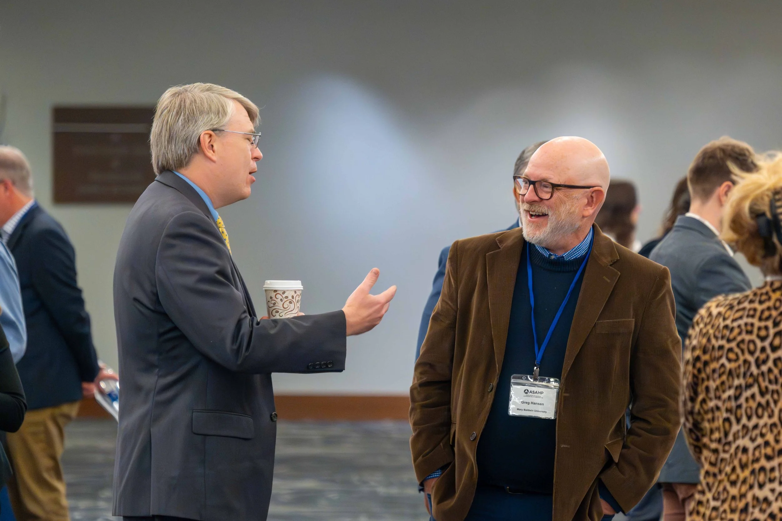 Two men talking at a professional event. One man is wearing a gray suit and yellow tie, holding a coffee cup, and gesturing with his hand. The other man is wearing a brown blazer, glasses, and a name badge, smiling while listening. Several other peop