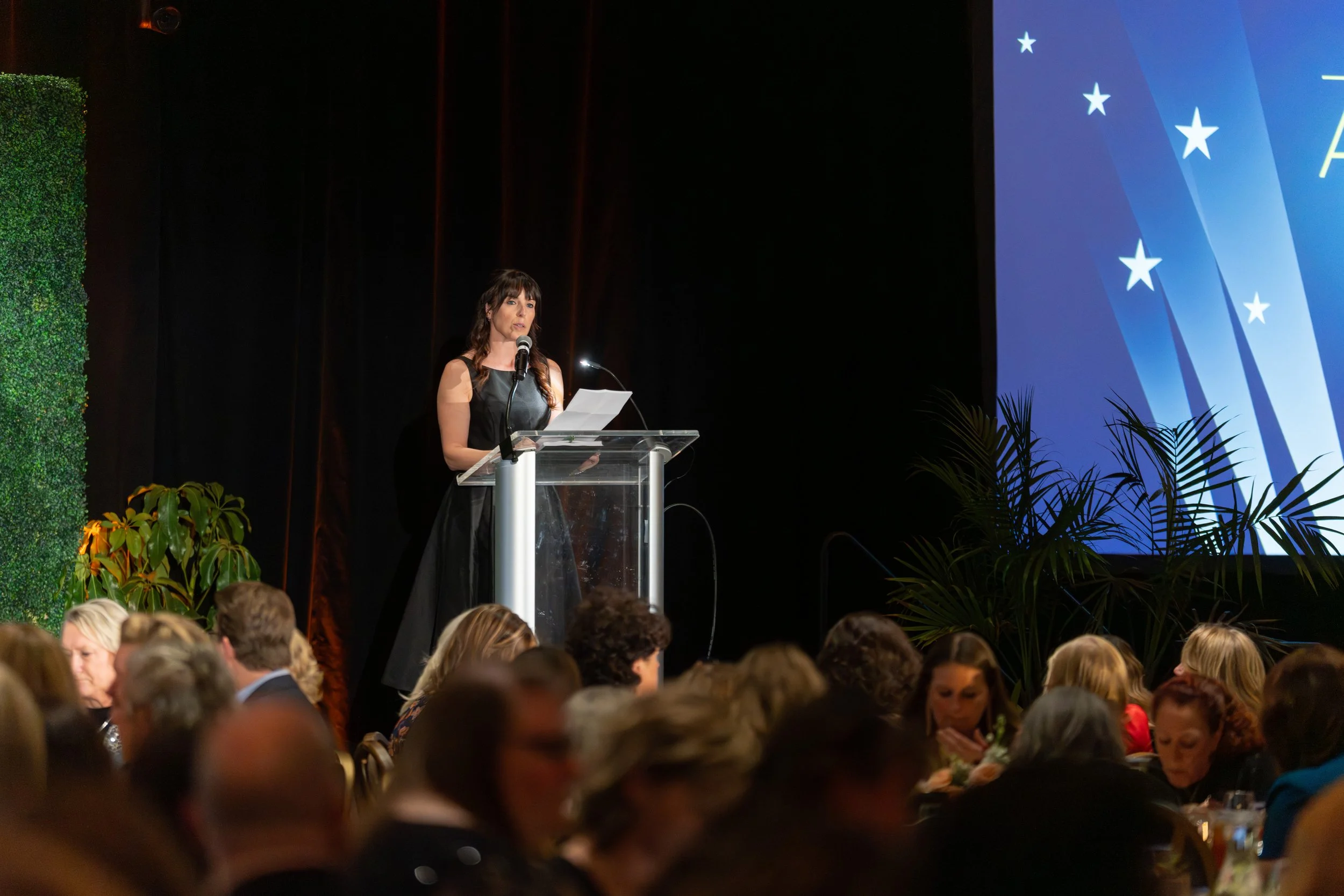 A woman in a black dress speaking at a podium during a formal event, with an audience seated in front of her and a large screen displaying a blue background with white stars on her right.