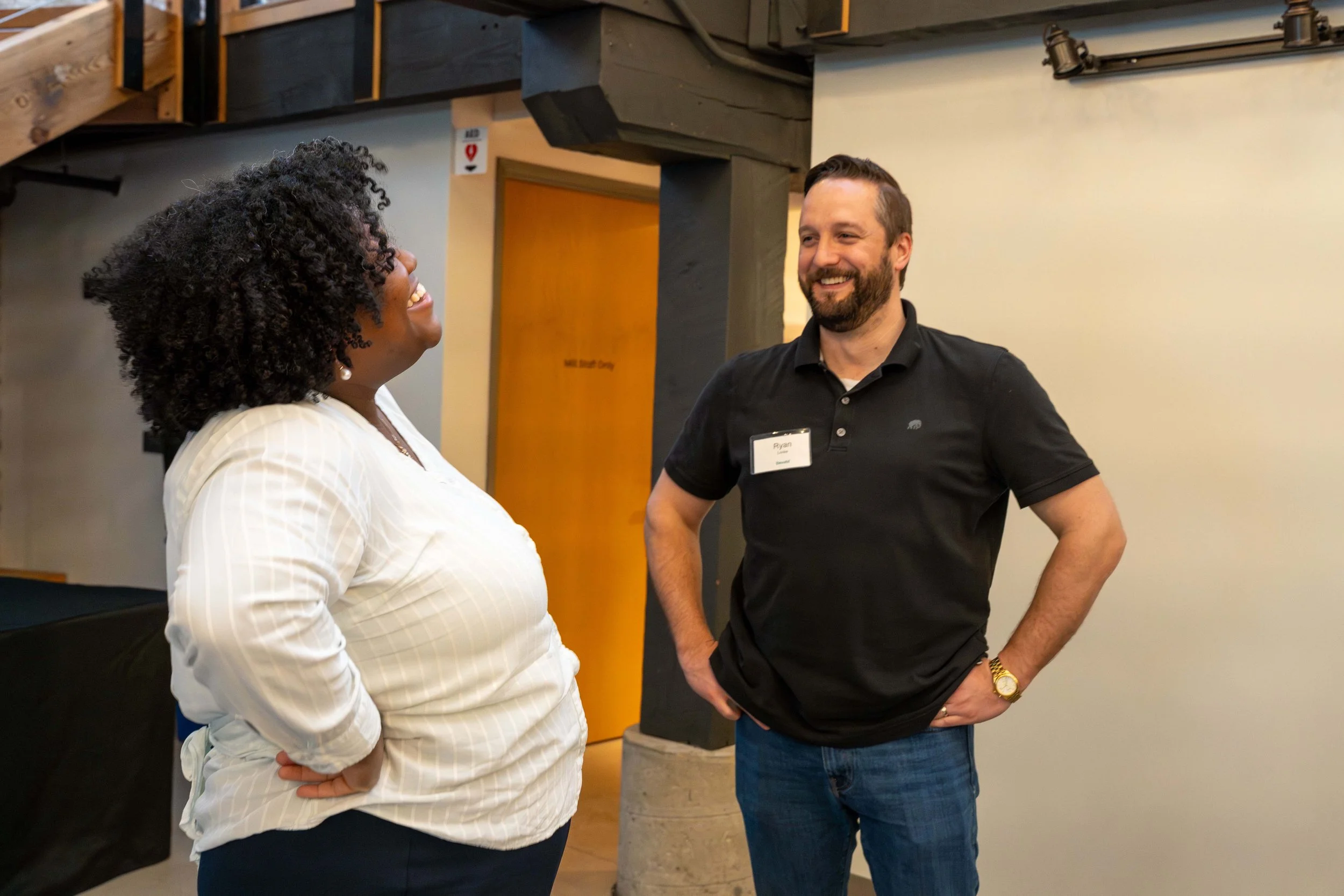 A woman and a man are smiling and having a conversation in an indoor setting. The woman has curly hair and is wearing a white blouse, while the man has a beard and is wearing a black polo shirt and jeans.