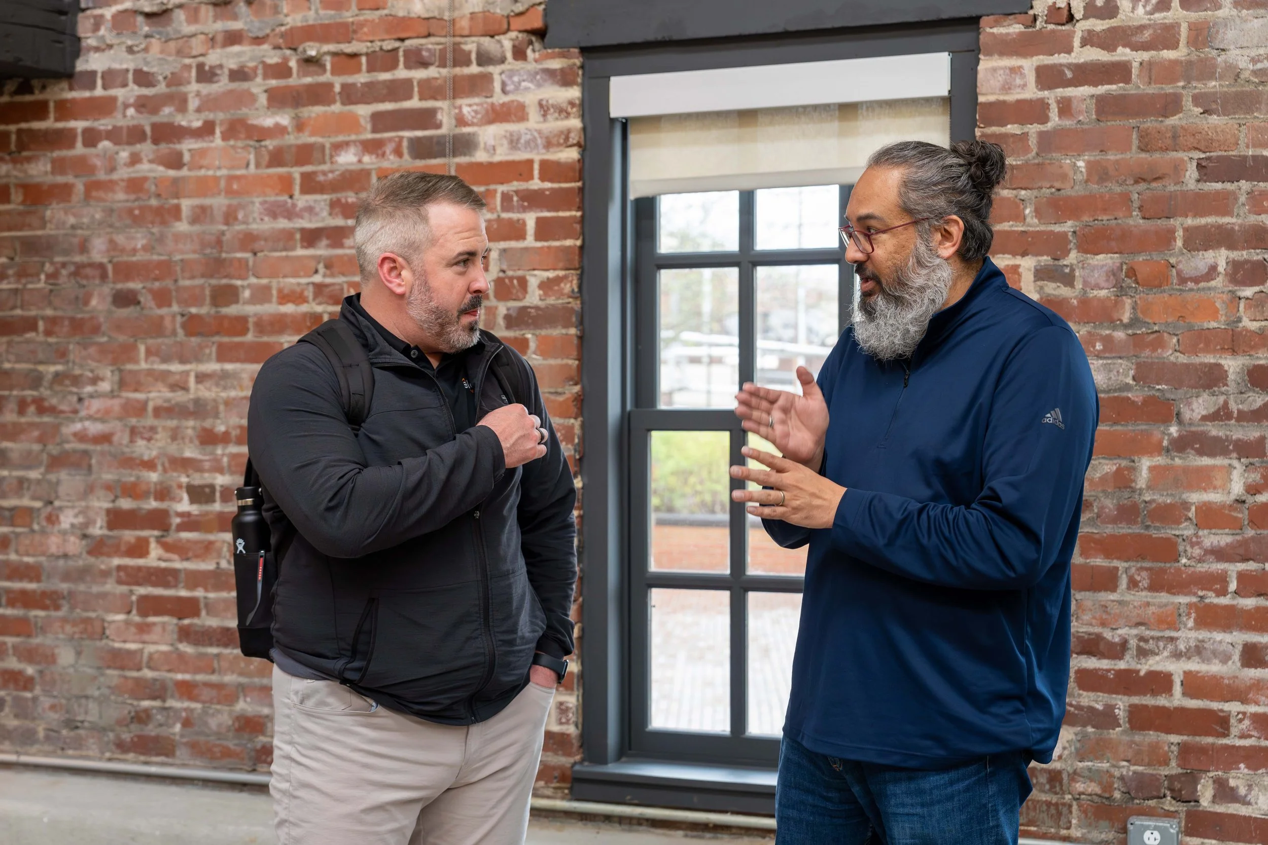 Two men having a conversation indoors against a brick wall. One man is wearing a black jacket with a backpack, and the other is wearing a blue jacket and glasses, with a gray beard and hair in a bun.