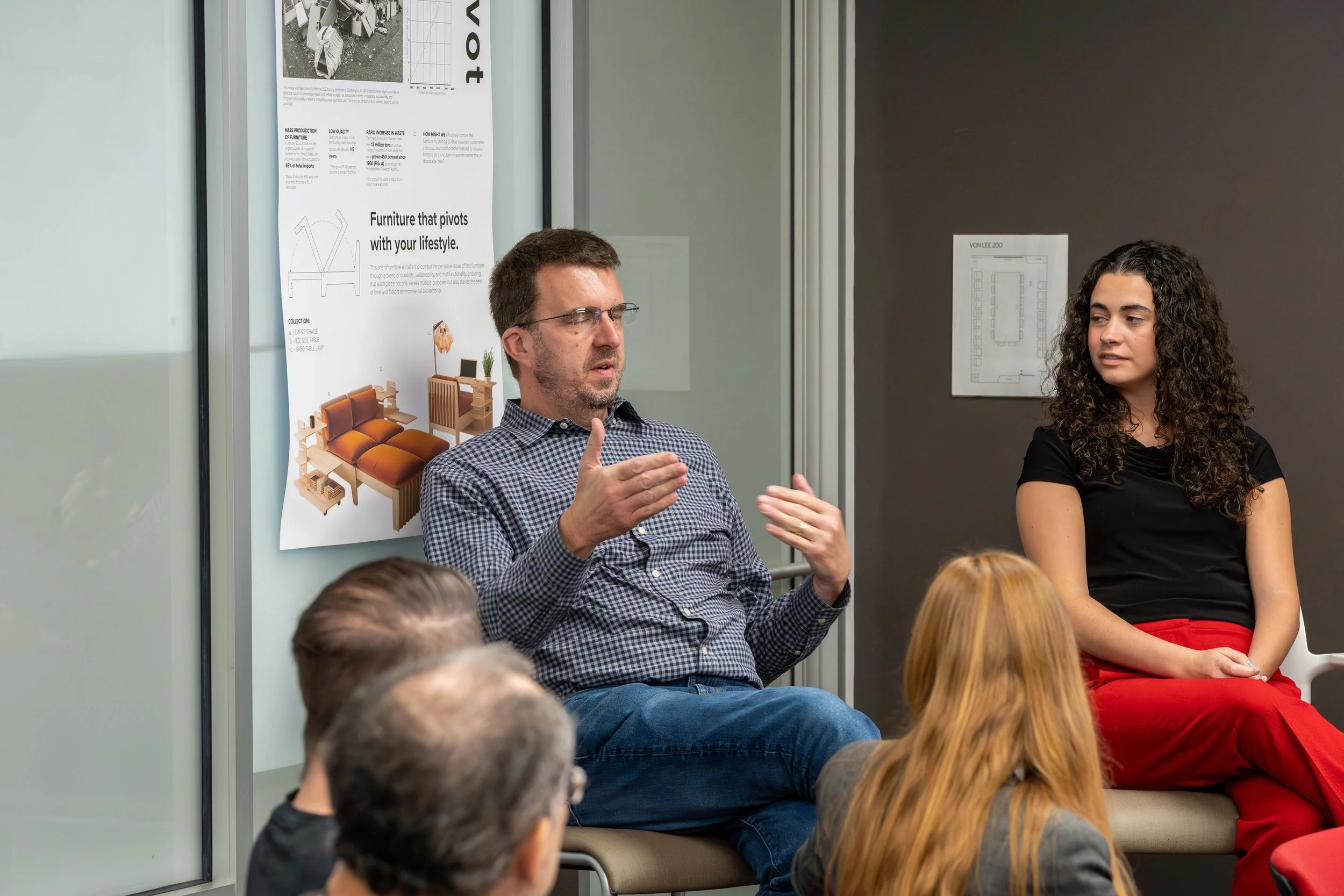 A man with glasses and a checkered shirt speaking in a discussion, with a woman in a black top and red pants sitting next to him, and several people in the audience listening.