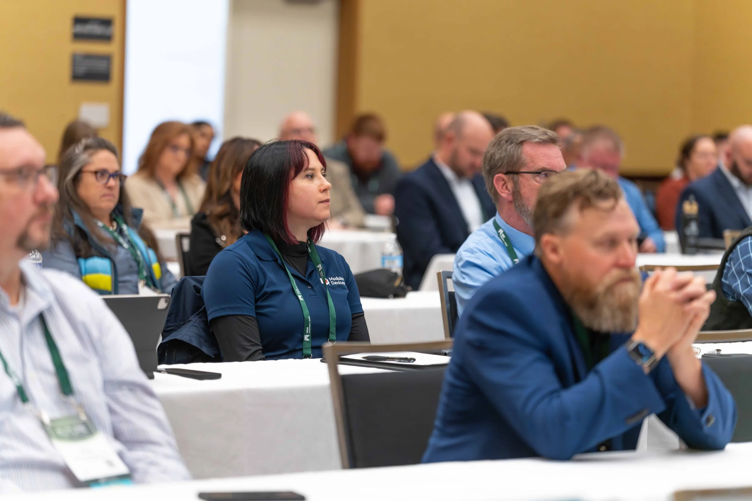 Conference attendees sitting at tables, listening attentively, in a large room.