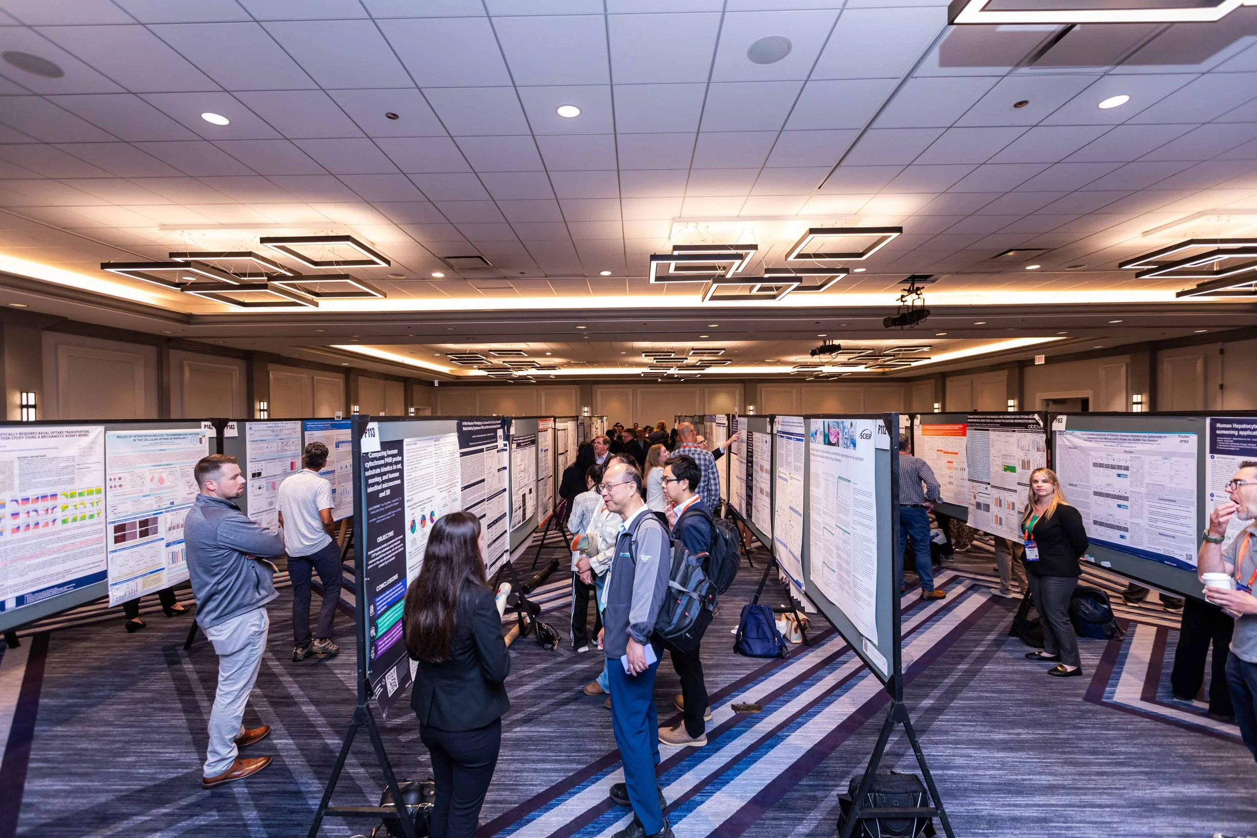 Conference attendees examining research posters in a large, well-lit conference room.
