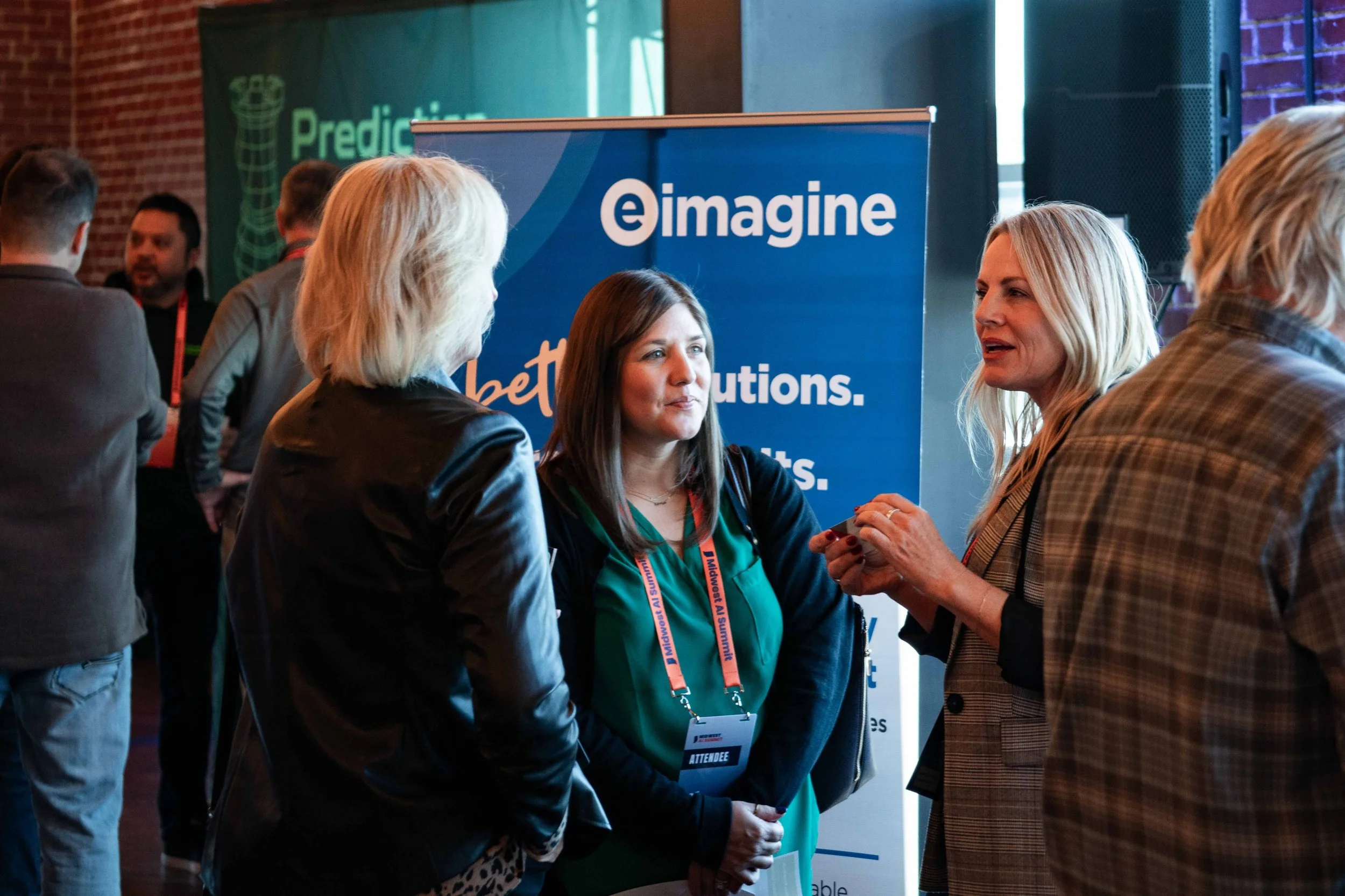 Three women engaged in conversation at a conference or networking event, with a blue banner behind them that reads 'eimagine' and has text about solutions and innovations. Other attendees are visible in the background.