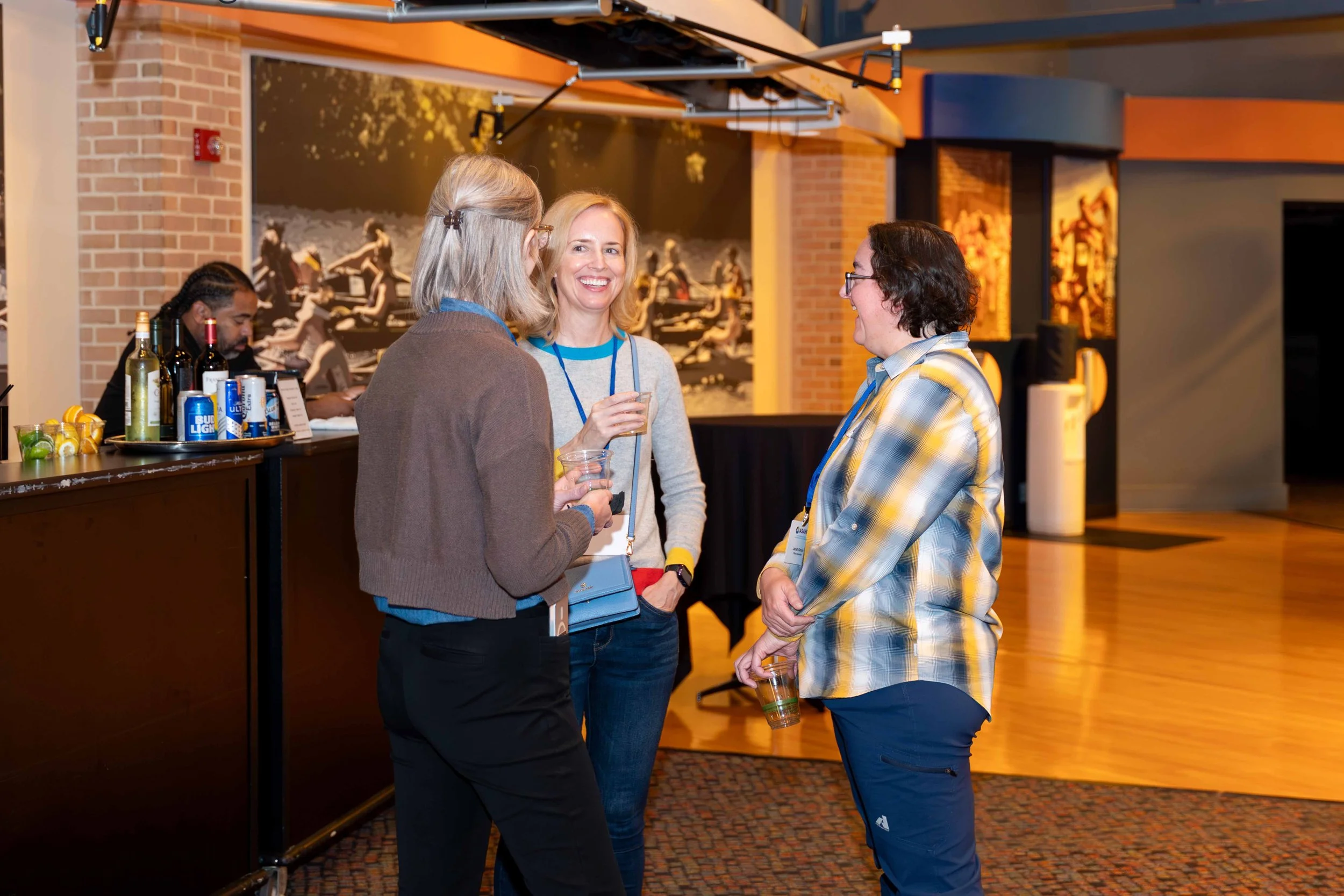 Three women are talking and smiling at a social event, with one holding a drink, in a room with a bar and a wall mural in the background.