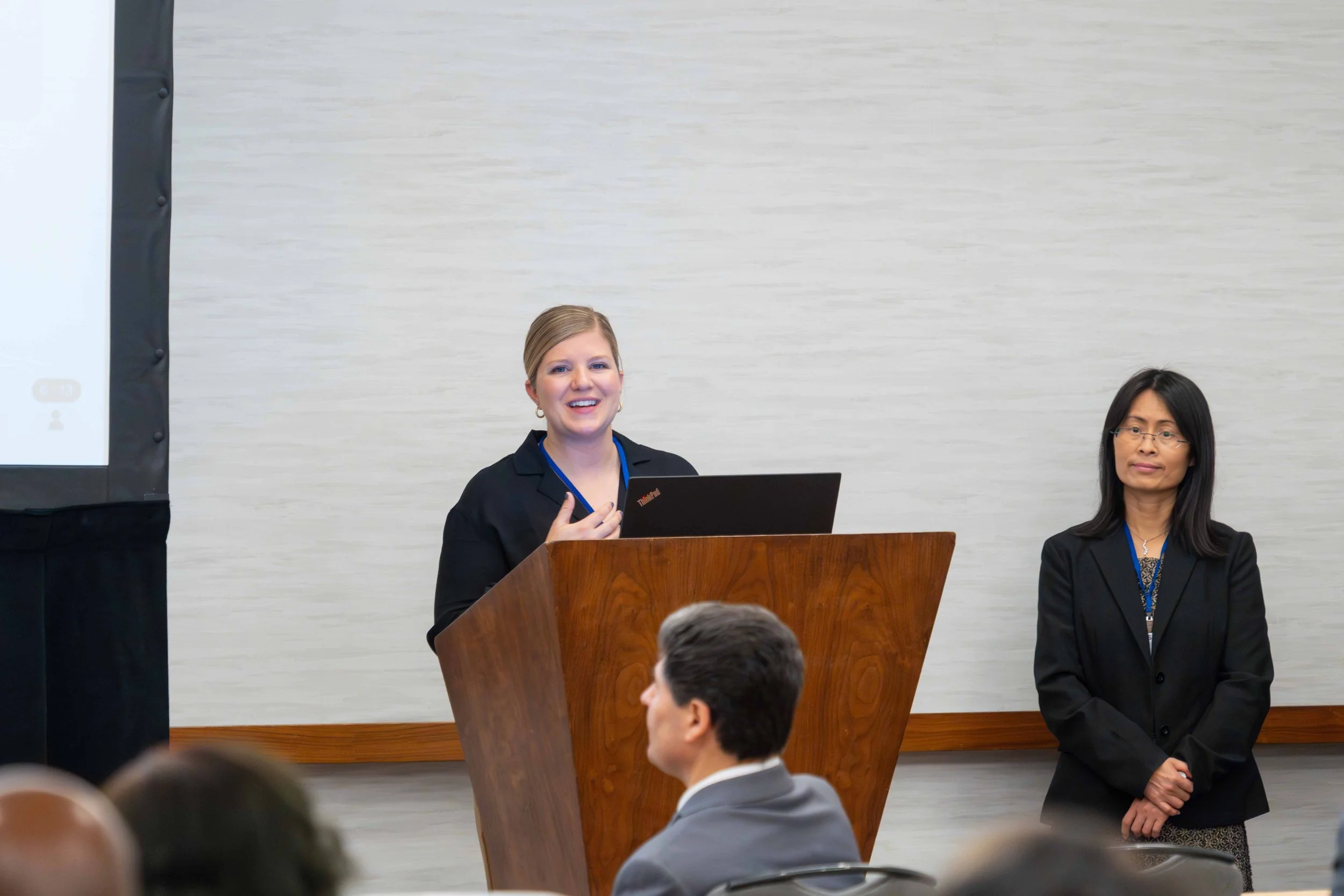 A woman with blonde hair speaking at a podium with a laptop, and another woman with black hair and glasses standing beside her, during a presentation or conference.