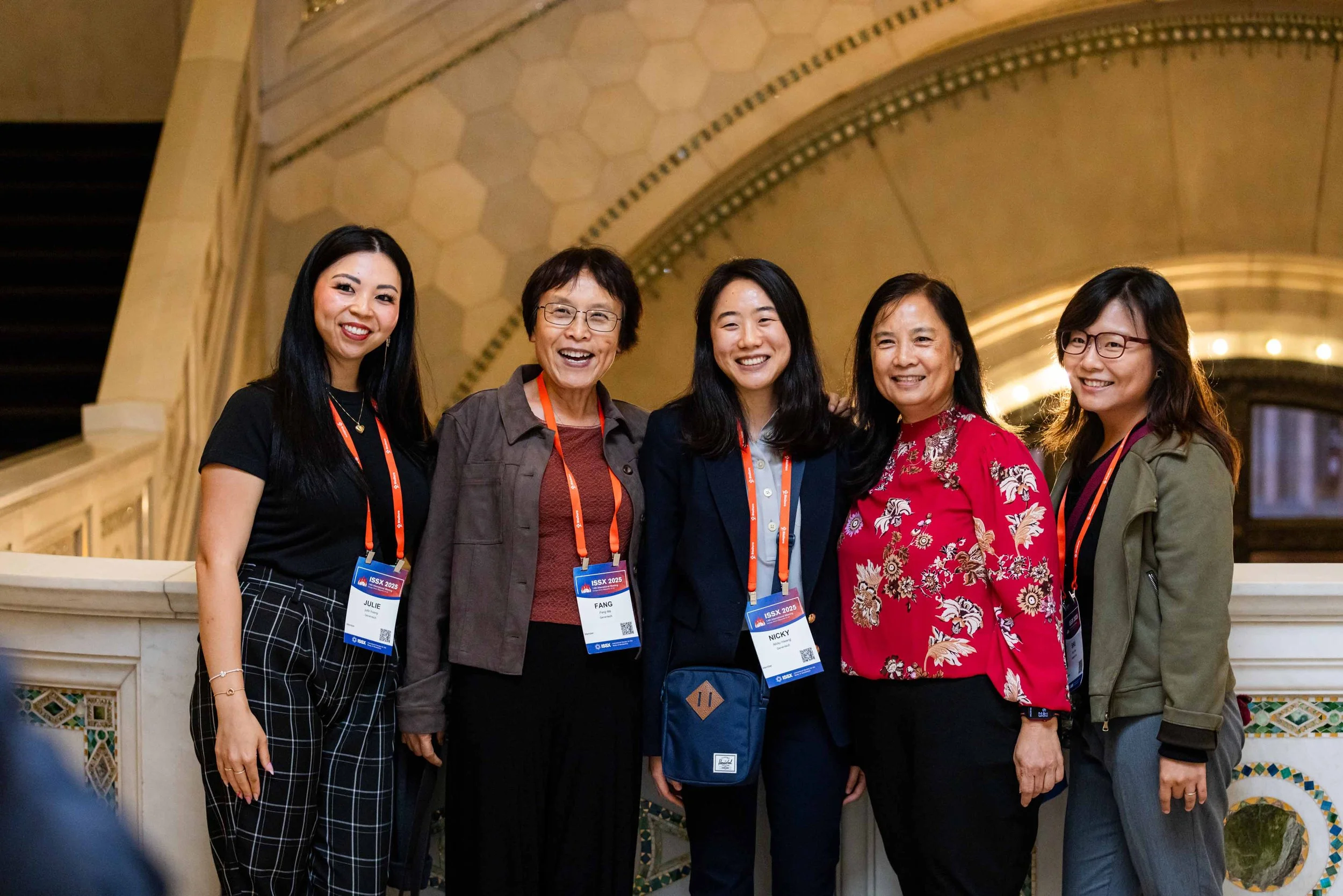 Five women standing together, smiling at an indoor event, wearing name badges, with a decorative arch and mosaic wall in the background.