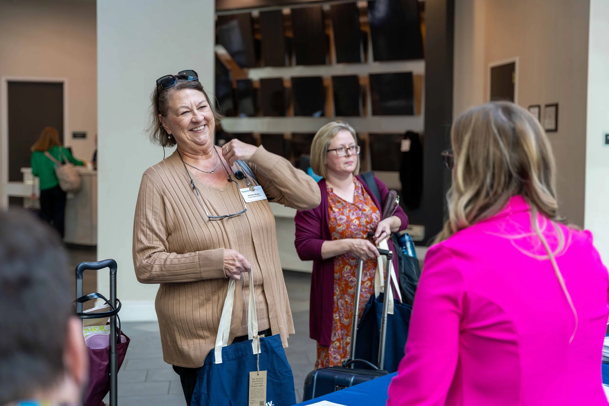 A woman in a beige sweater holding a tote bag and smiling at a registration table in a busy indoor setting, with other women and luggage around.