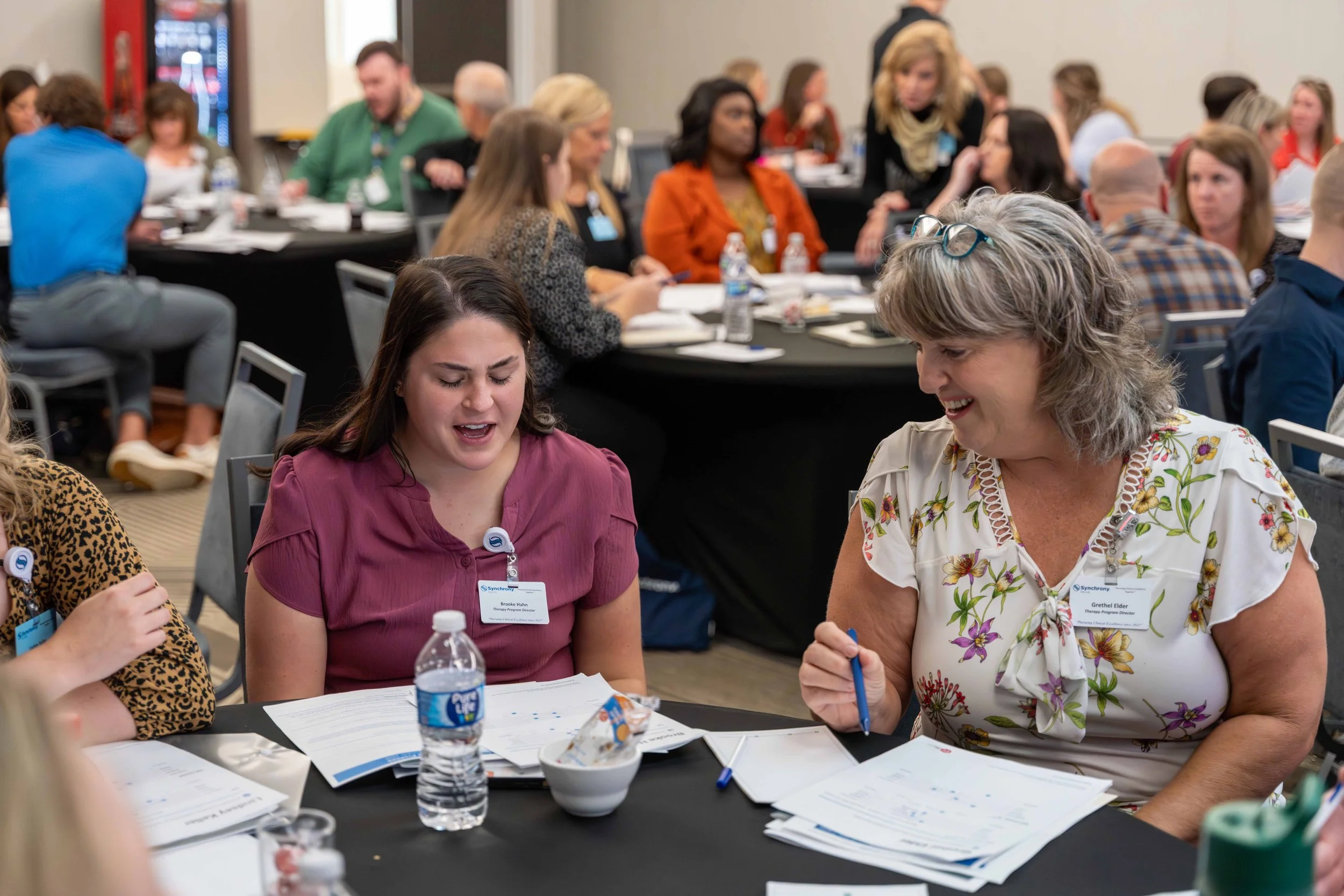 Two women are seated at a conference table, engaged in conversation and smiling, with documents, pens, and water bottles on the table.