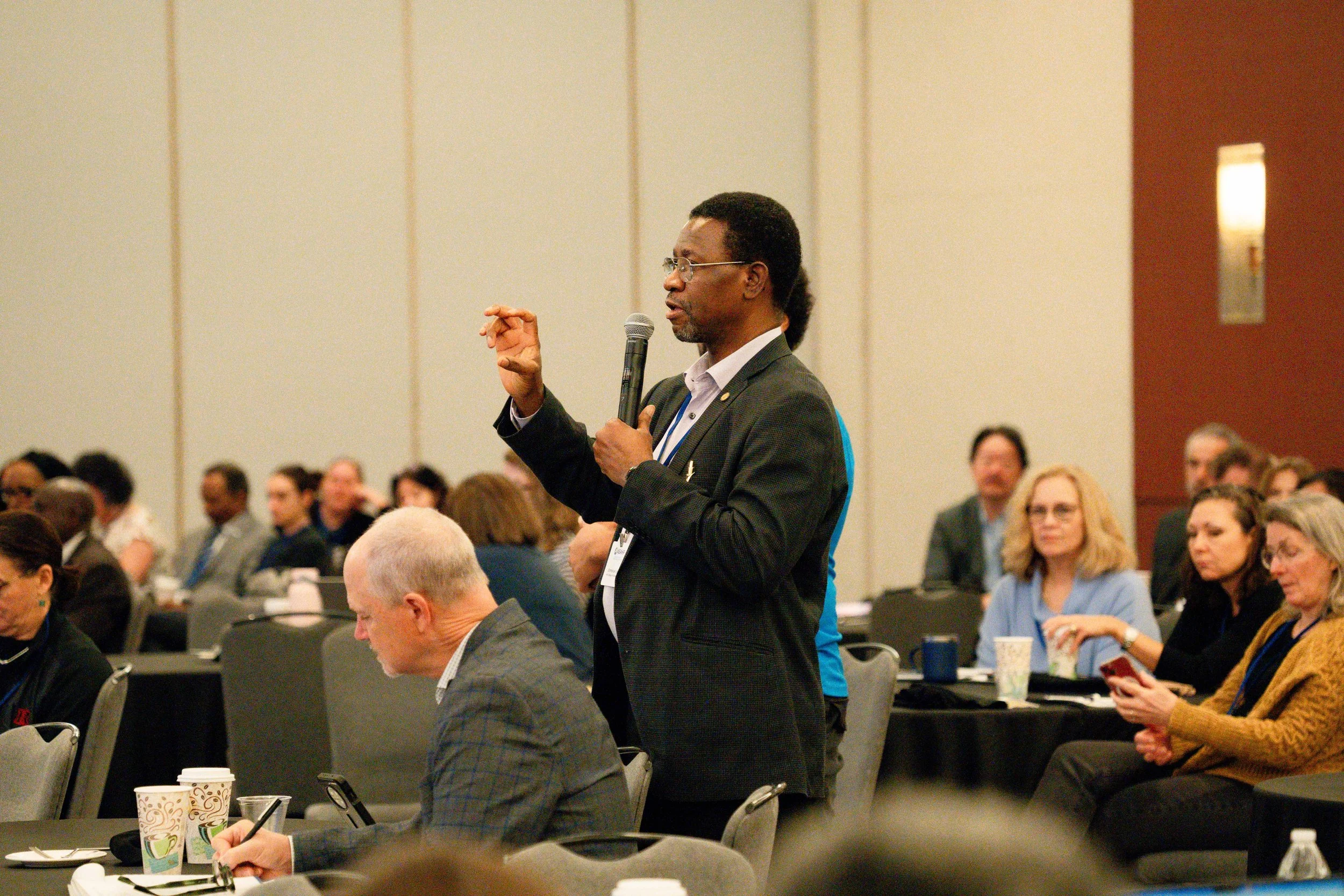 A man in a dark suit holding a microphone and speaking at a conference with seated attendees listening in the background.