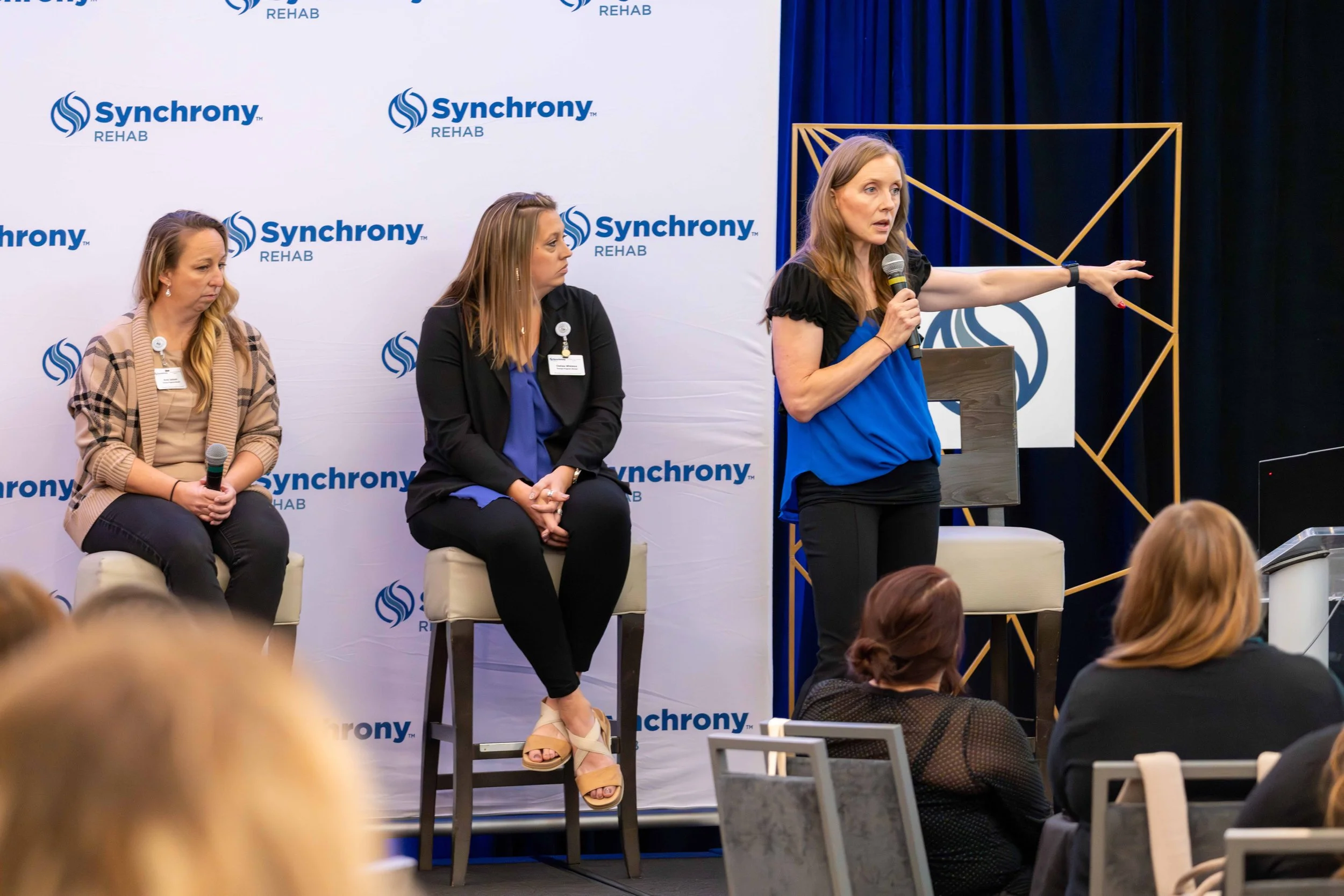 Women participating in a panel discussion at an event with a backdrop displaying the 'Synchrony Rehab' logo. The woman on the right is standing, holding a microphone, and pointing toward the right. Two women are seated on stools to her left, listenin