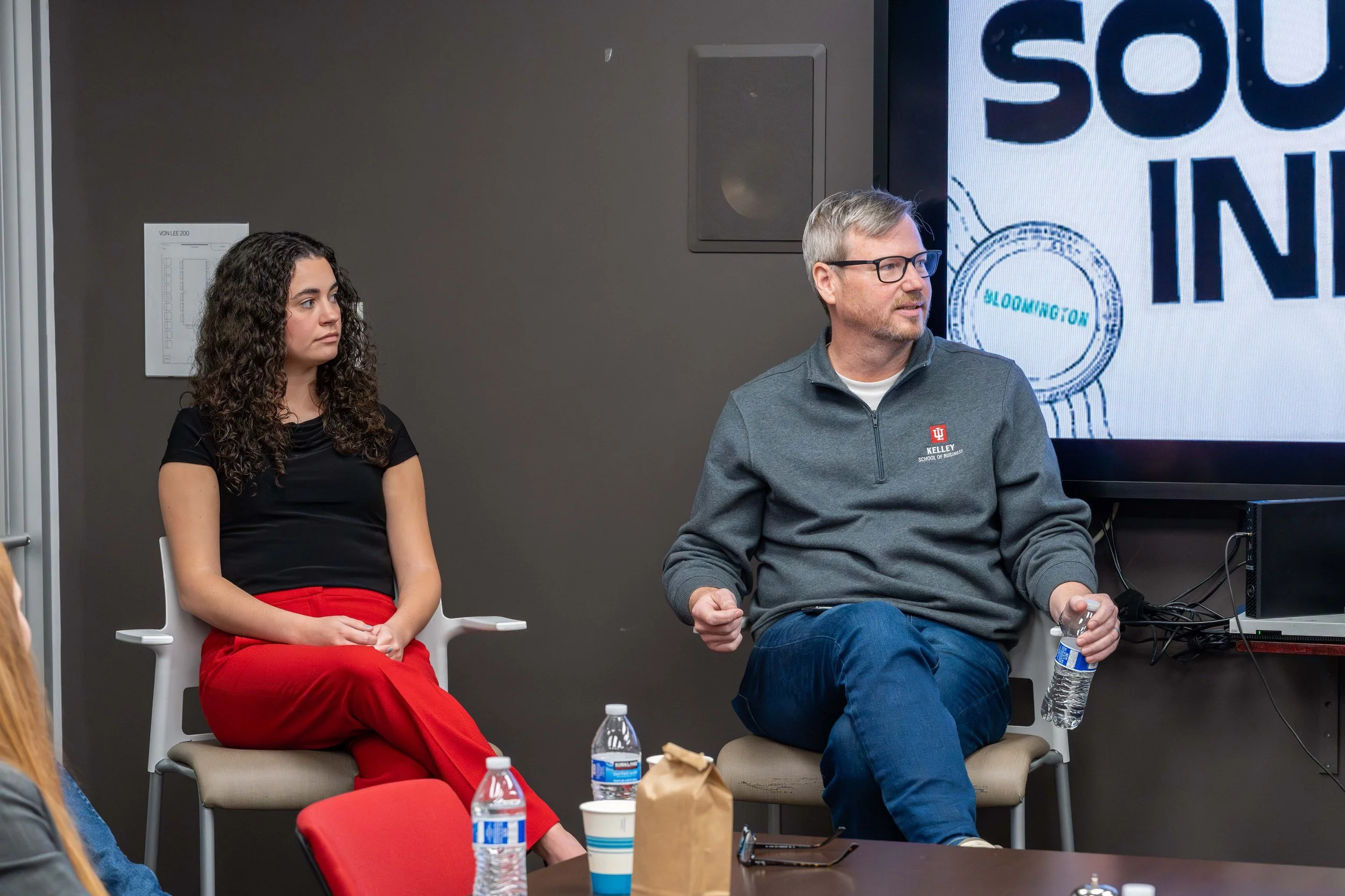 Two people sitting in chairs during a discussion or presentation. The woman on the left has curly dark hair and is wearing a black top and red pants. The man on the right has short gray hair, glasses, and is wearing a gray sweatshirt with a logo, hol