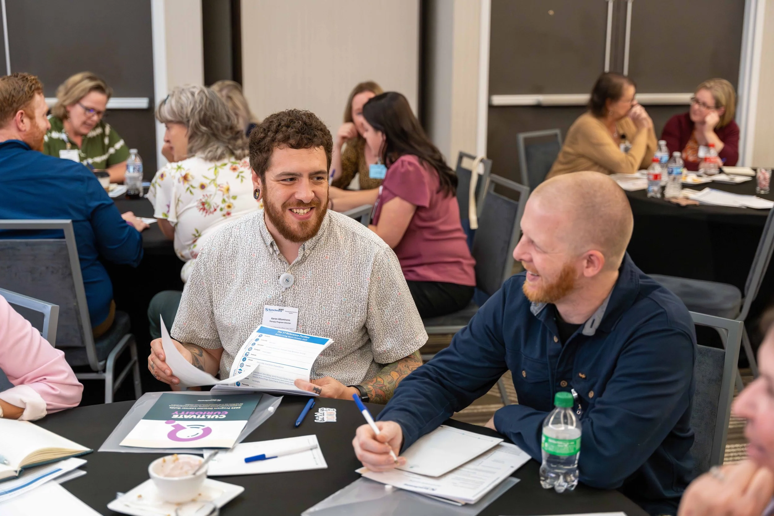 Two men seated at a conference table smiling and talking, with other attendees in the background, some talking and others listening, in a conference room setting with notebooks, papers, and water bottles on the table.