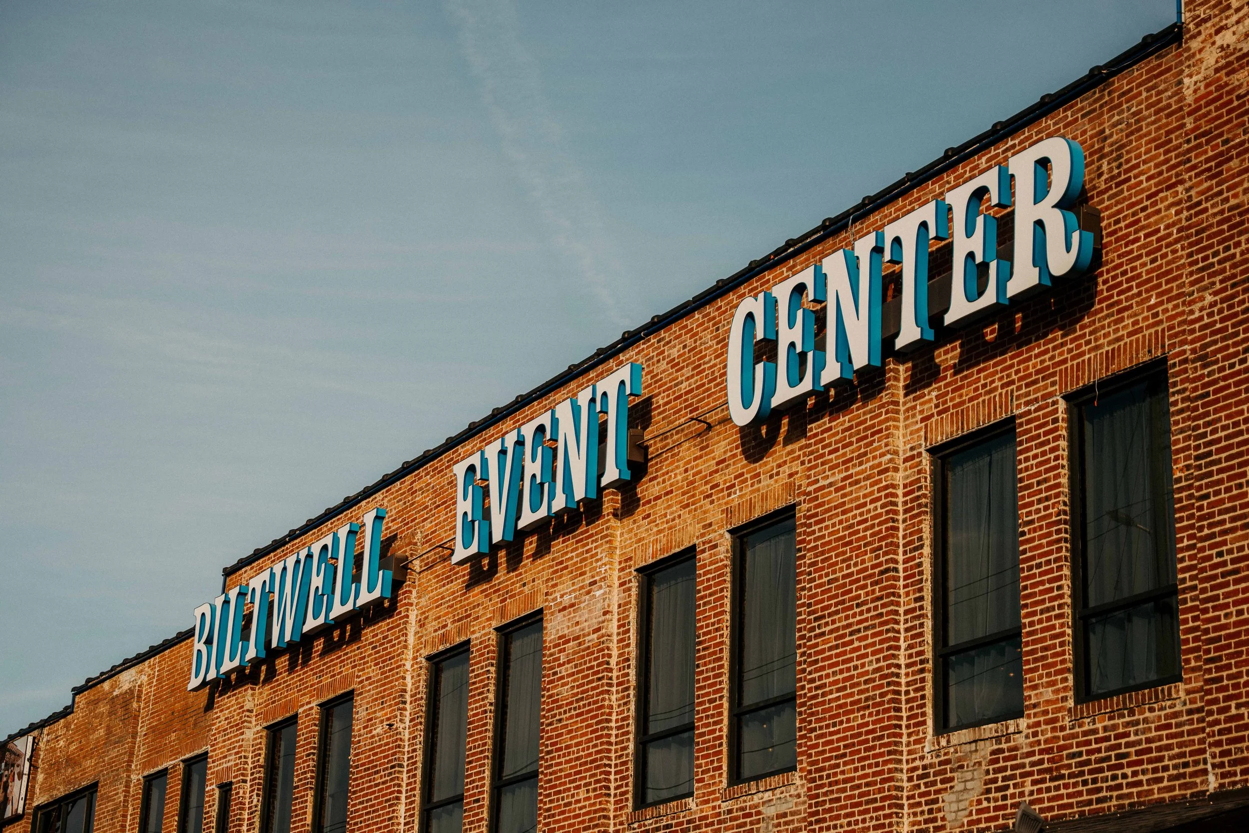 Brick building with large blue and white sign reading "BILTWELL EVENT CENTER" on the upper facade, multiple black window frames, and a clear sky in the background.