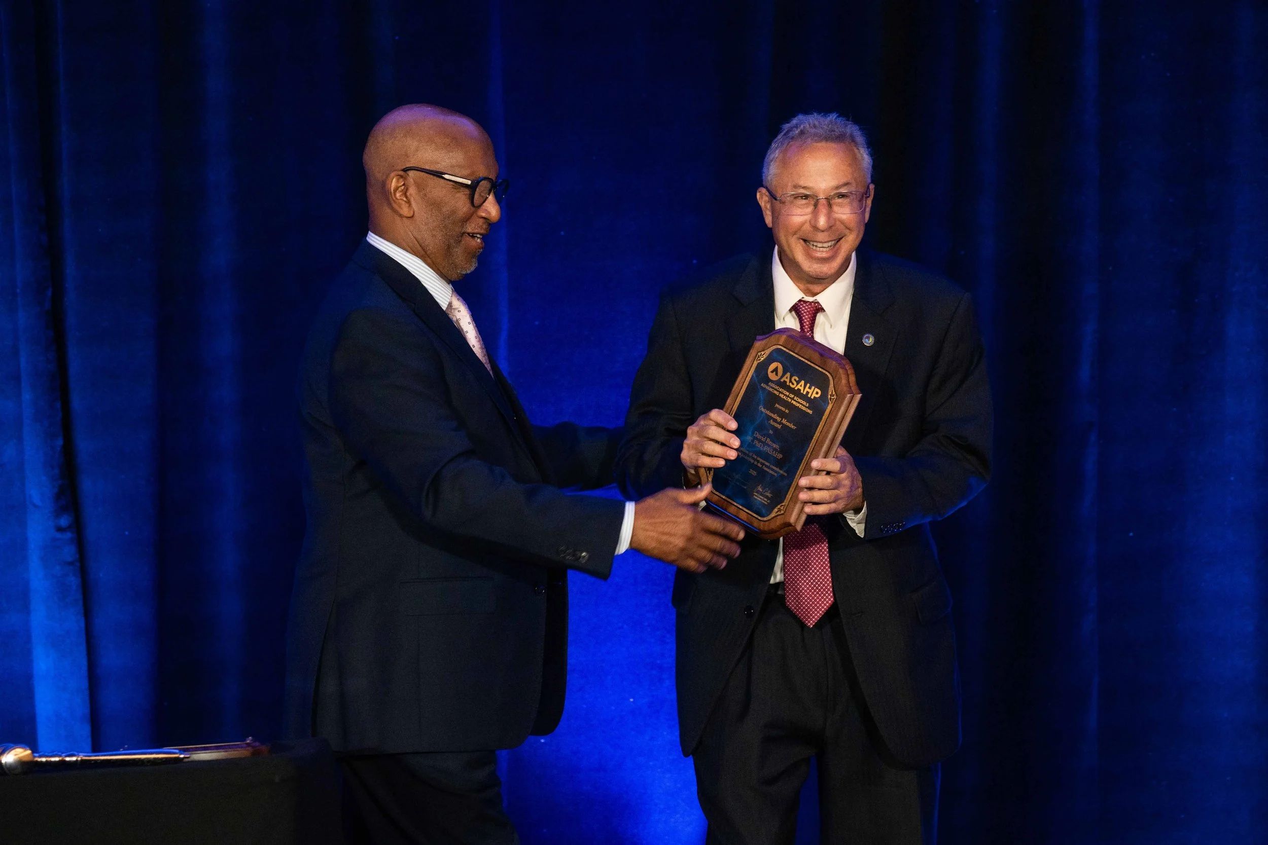 Two men in suits on stage, one presenting an award plaque to the other, with dark blue curtains in the background.