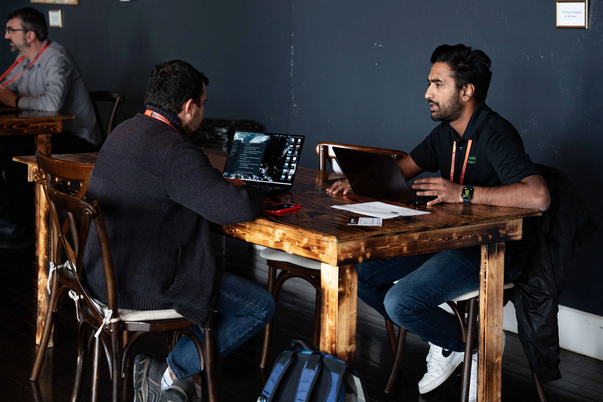 Two men engaged in a discussion at a wooden table, using laptops, in a casual indoor setting.