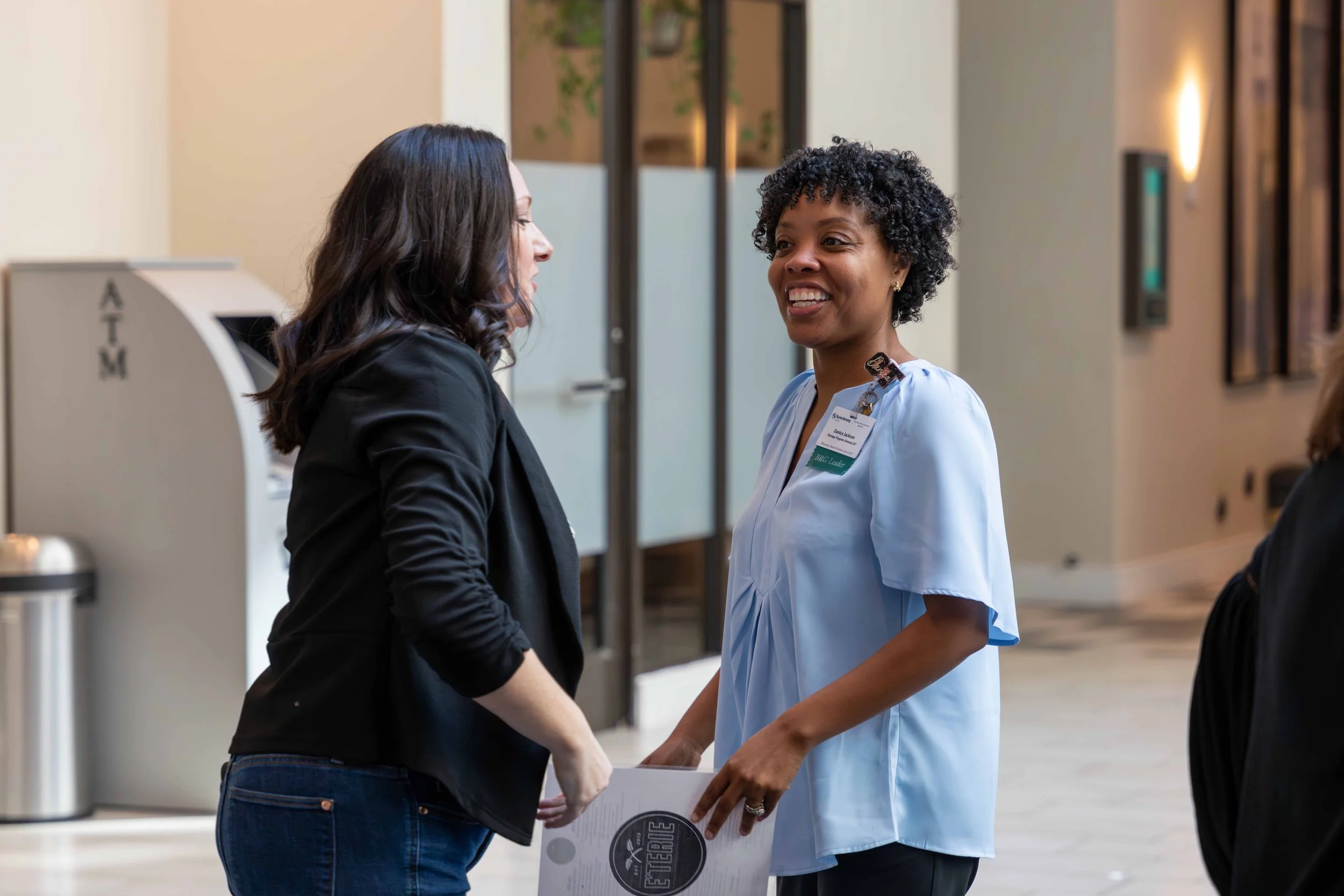 Two women engaged in conversation, one wearing a black blazer and the other in a light blue medical uniform with a name tag, in a professional indoor setting.
